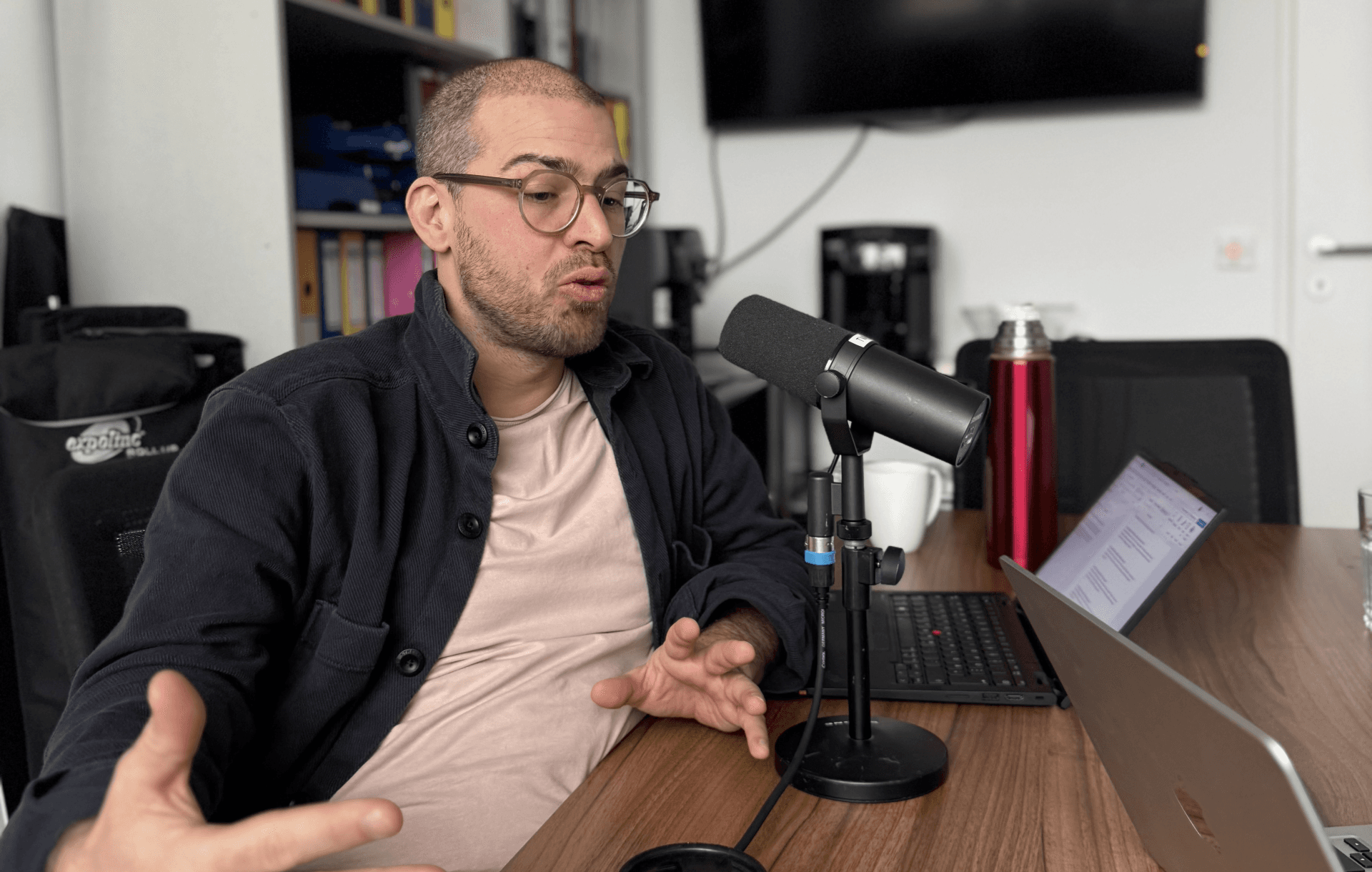 Ein Mann mit Brille und dunkler Jacke spricht in ein Mikrofon an einem Schreibtisch mit einem Laptop, einer Tasse und einem roten Becher. Im Hintergrund sind Regale mit Aktenordnern und ein großer, an der Wand montierter Fernseher zu sehen.