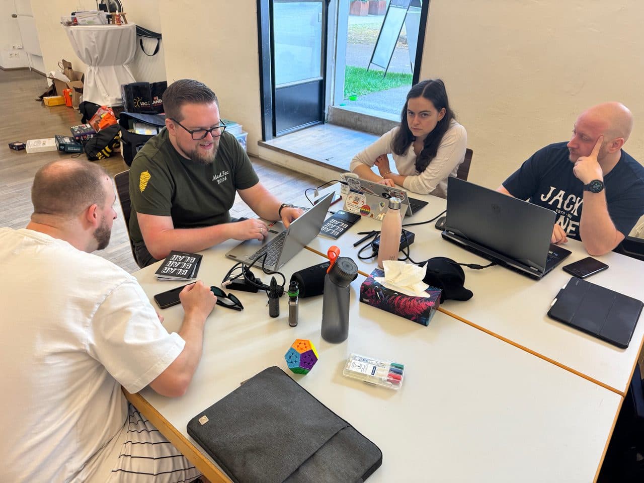 Vier Erwachsene sitzen an einem Tisch mit Laptops, arbeiten und diskutieren etwas. Auf dem Tisch stehen Wasserflaschen, ein Federmäppchen, ein Zauberwürfel und andere Schreibwaren. Hinter ihnen ist eine Tür nach draußen geöffnet.