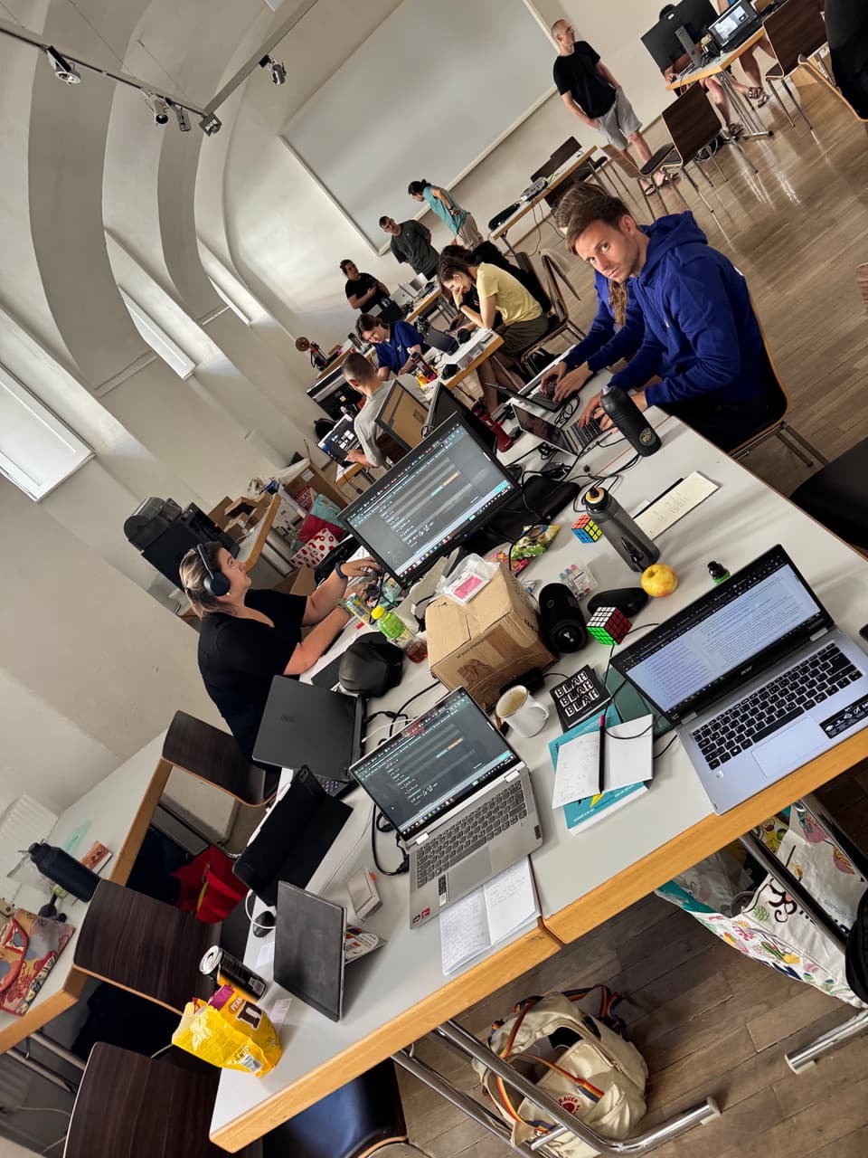 Menschen arbeiten an Laptops an einem gemeinsamen Arbeitsplatz, an dem Papiere, Snacks und Getränke verstreut sind. Einige tippen, andere unterhalten sich, und im Hintergrund sind weitere Personen zu sehen. Der Raum hat hohe Decken und große Fenster.