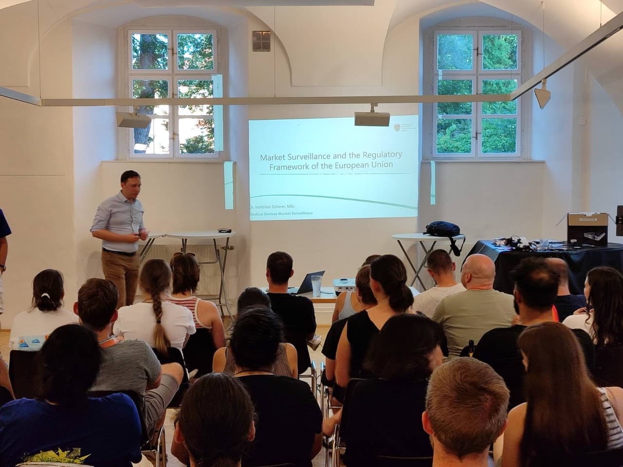 Ein Mann steht vor einem sitzenden Publikum und hält einen Vortrag mit dem Titel 'Market Surveillance and the Regulatory Framework of the European Union' (Marktüberwachung und der Rechtsrahmen der Europäischen Union), der auf einem Projektionsbildschirm in einem Klassenzimmer mit großen Fenstern angezeigt wird.