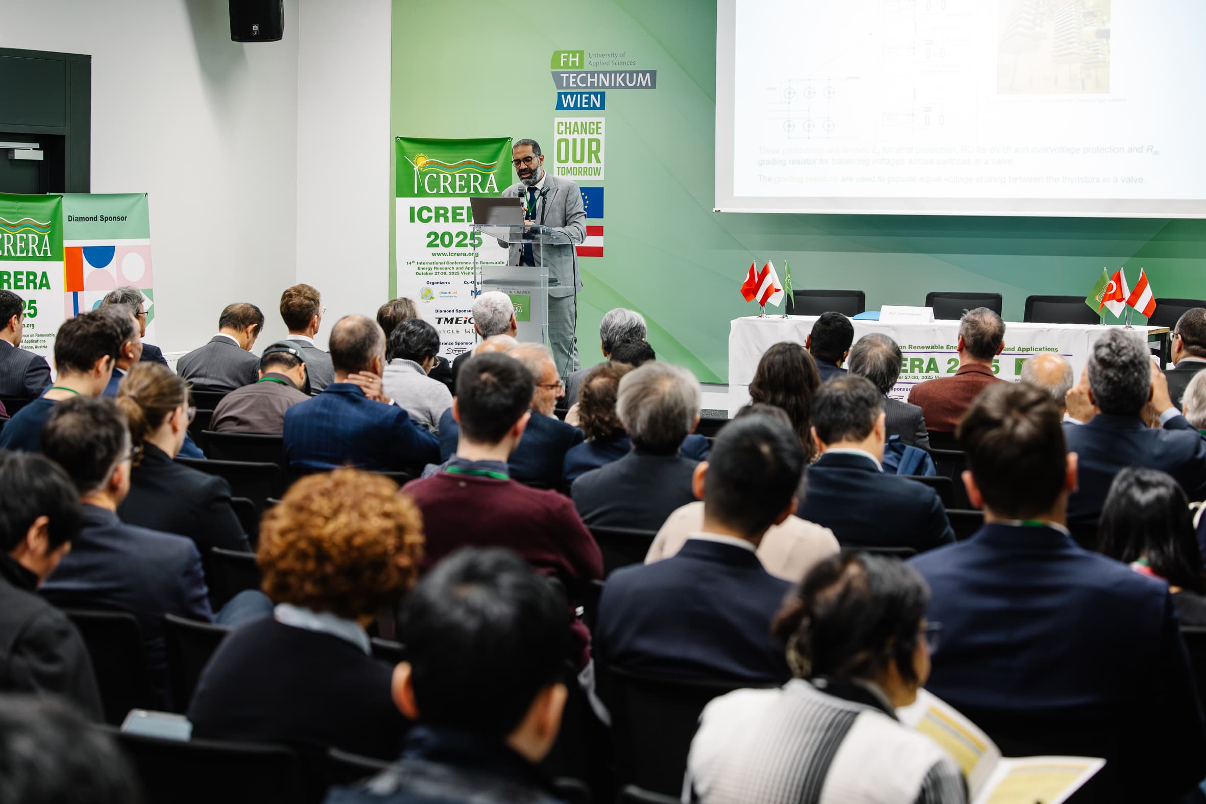 Ein Redner spricht auf einer Konferenz in einem modernen Hörsaal zu den Zuhörern. Die Zuhörer sitzen mit Blick auf die Bühne, auf der Präsentationsfolien und Schilder für ICRERA 2025 zu sehen sind. Auf dem Tisch sind Nationalflaggen angebracht.