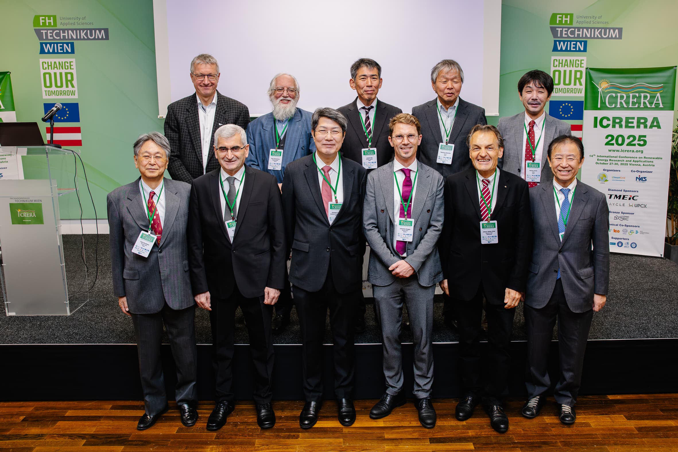 Eine Gruppe von elf Männern in formeller Kleidung, die Konferenzabzeichen tragen, steht gemeinsam auf einer Bühne bei einer Fachveranstaltung. Hinter ihnen sind Banner und ein Podium mit den Logos der FH Technikum Wien und ICRERA 2025 zu sehen.