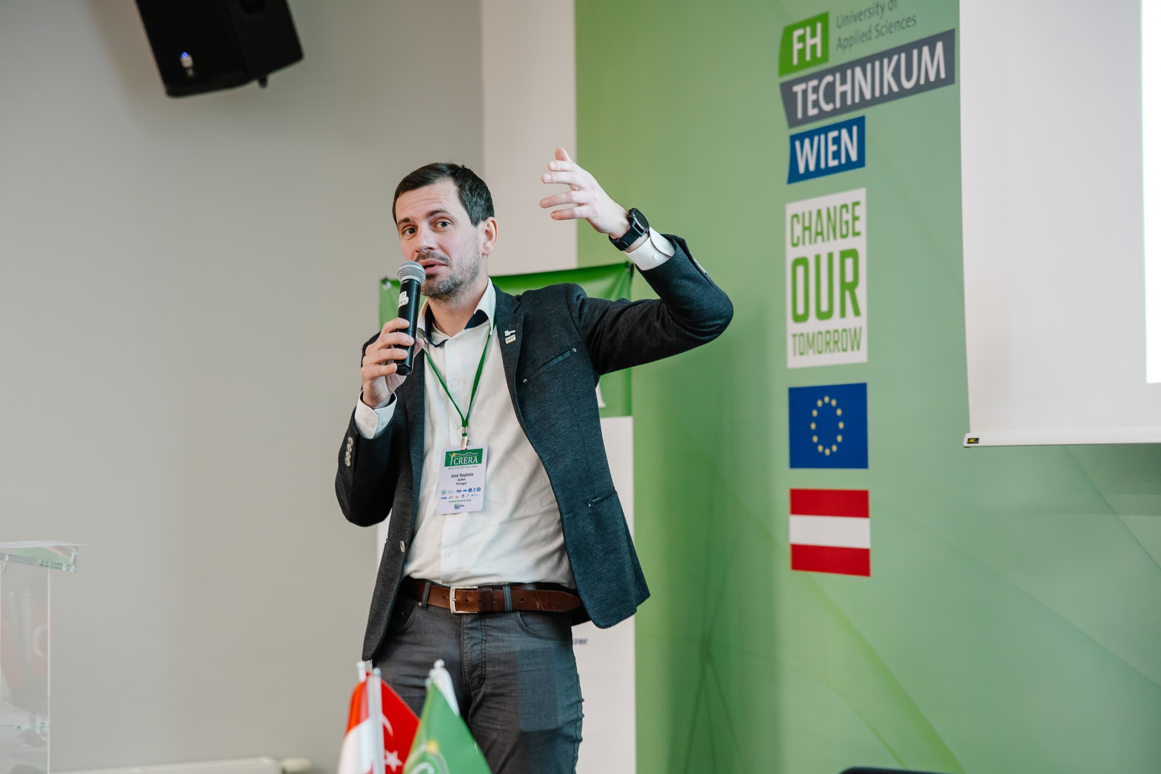Ein Mann im Anzug spricht in ein Mikrofon und gestikuliert mit einer Hand während einer Präsentation an der FH Technikum Wien. Im Hintergrund sind Fahnen und ein Change Our Tomorrow-Schild zu sehen.