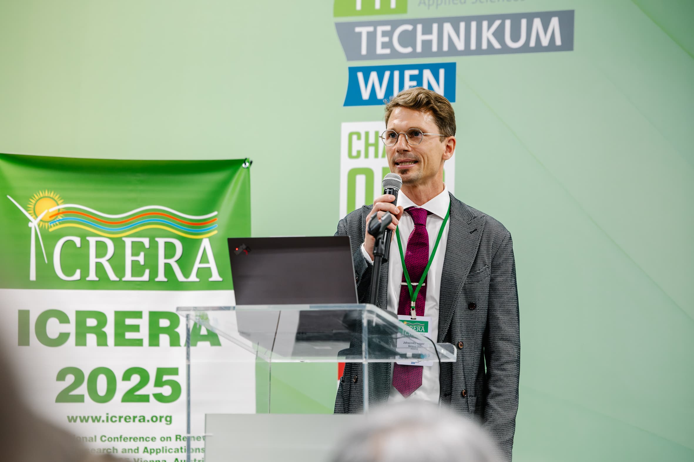 Ein Mann in Anzug und Krawatte spricht an einem Podium mit einem Mikrofon. Hinter ihm sind Konferenzbanner zu sehen, darunter eines mit der Aufschrift ICRERA 2025 und ein weiteres mit TECHNIKUM WIEN. Ein Laptop steht auf dem Podium.