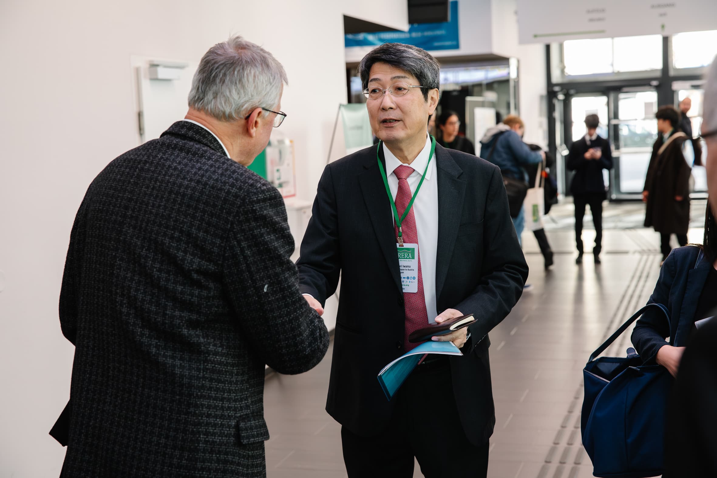 Zwei Männer in Geschäftskleidung schütteln sich die Hände und unterhalten sich in einem modernen Flur während einer Konferenz oder Veranstaltung. Im Hintergrund sind andere Personen zu sehen, die sich unterhalten oder spazieren gehen.