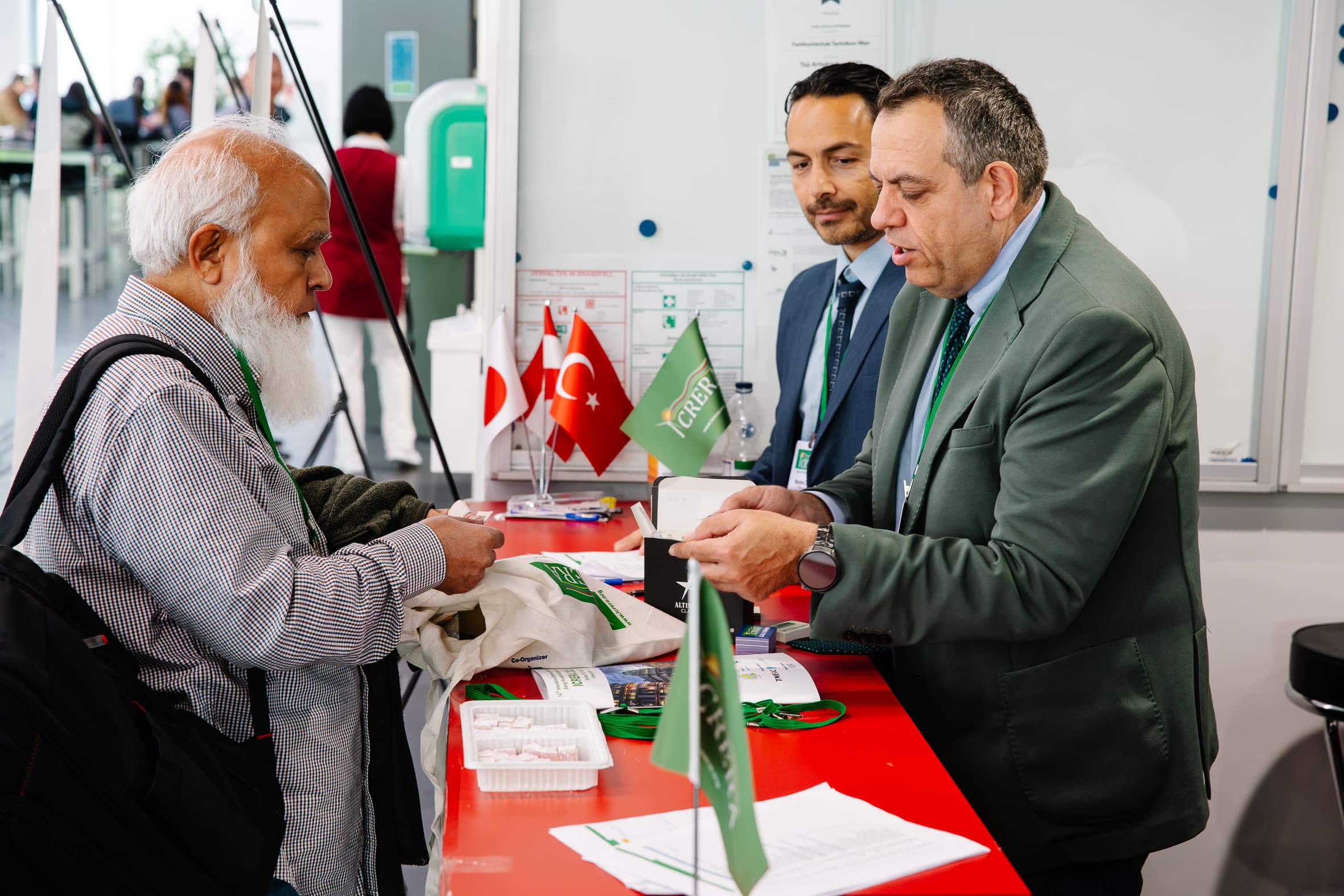 Ein Mann an einem Schalter spricht mit zwei Mitarbeitern in Anzügen. Auf dem Tresen liegen türkische und japanische Flaggen, Broschüren und grüne Banner. Eine weitere Person steht im Hintergrund.