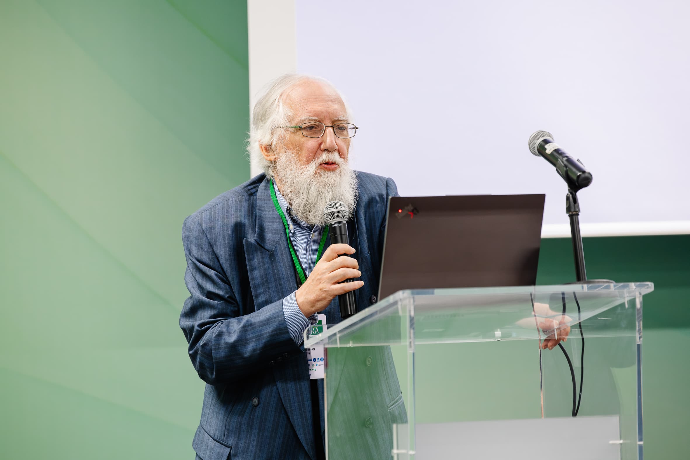 Ein älterer Mann mit weißem Bart und Brille spricht in ein Mikrofon, während er an einem durchsichtigen Podium mit einem Laptop steht. Er trägt einen blauen Anzug und ein Namensschild vor einem grünen Hintergrund.