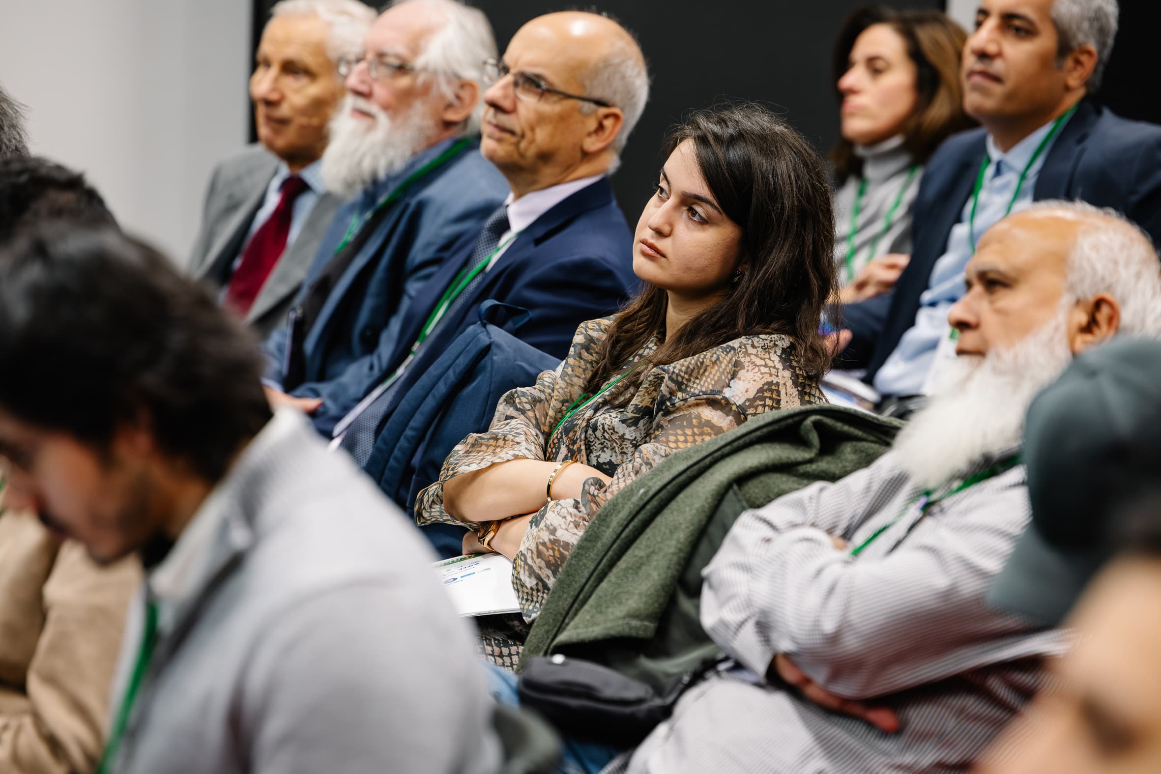 Eine Gruppe von Personen, Männer und Frauen unterschiedlichen Alters, sitzt eng beieinander in einem Konferenzraum und hört aufmerksam zu. Die meisten tragen Geschäftskleidung mit grünen Schlüsselbändern und einige haben einen nachdenklichen oder konzentrierten Gesichtsausdruck.