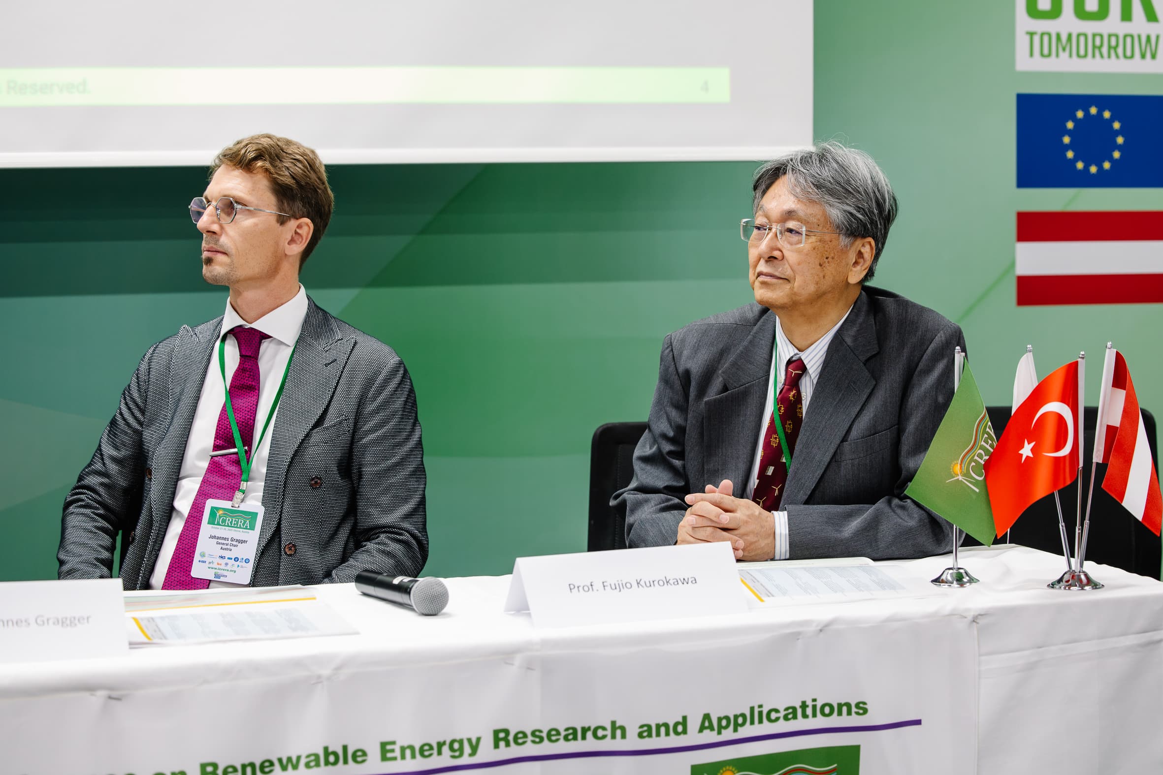 Zwei Männer in Anzügen sitzen an einem Konferenztisch mit kleinen Flaggen von Portugal, der Türkei, der EU und einer weiteren Flagge. Namensschilder weisen sie als Jens Gragger und Prof. Fujio Kurokawa aus. Im Hintergrund sind ein grünes Banner und ein Bildschirm zu sehen.