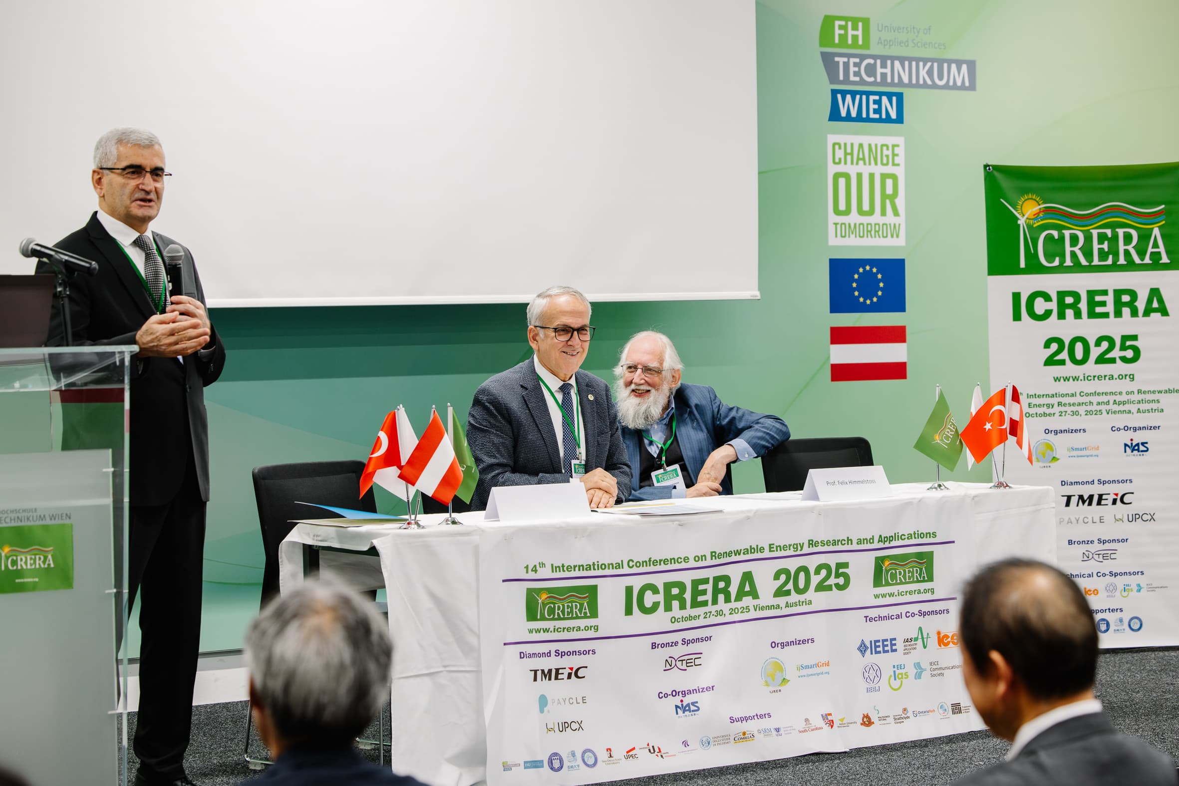 Drei Männer nehmen an einer Konferenz teil; einer spricht an einem Podium, während zwei an einem Tisch mit Mikrofonen und kleinen Fahnen sitzen. Die Kulisse und die Schilder werben für die Internationale Konferenz zur Forschung über erneuerbare Energien 2025.