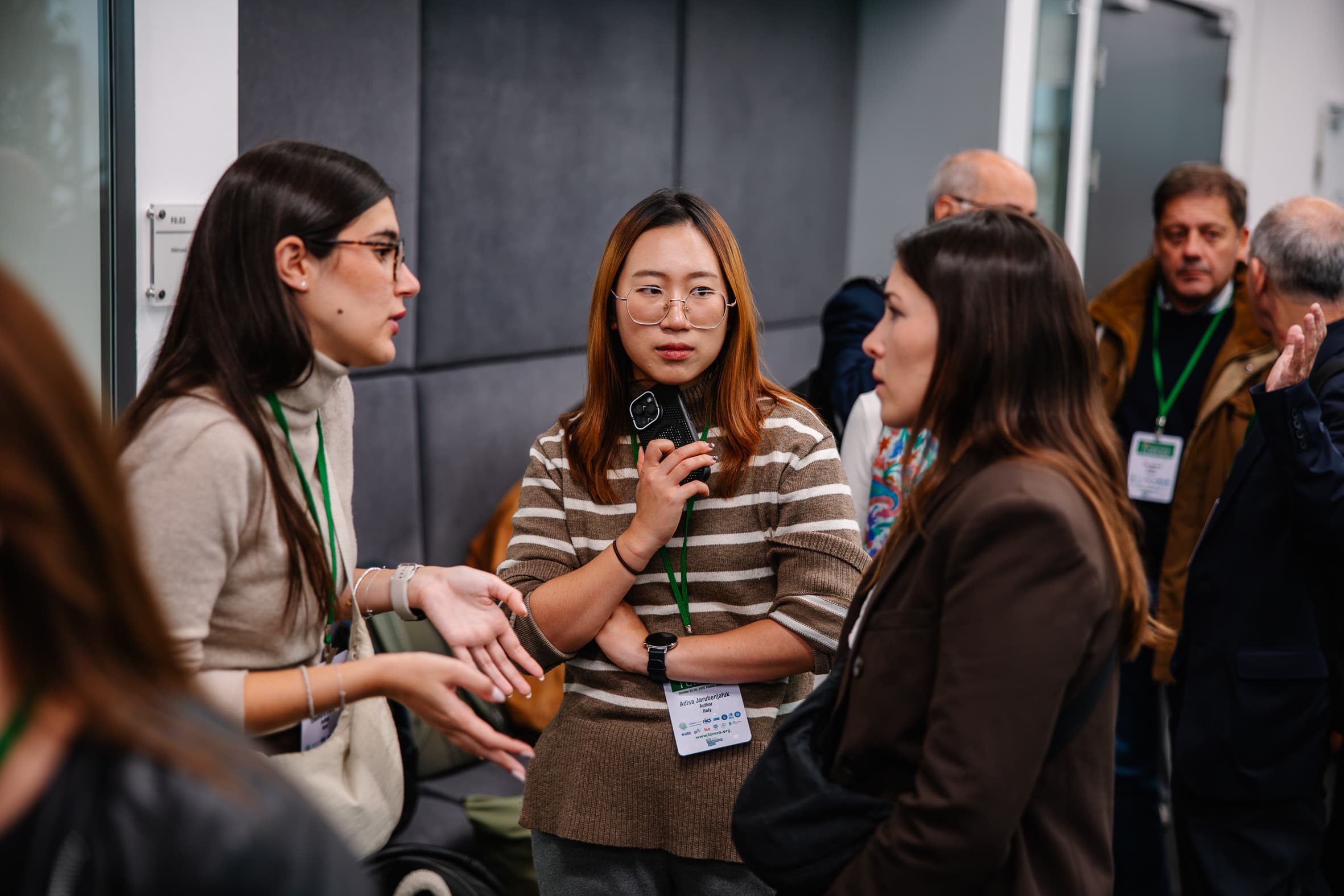 Drei Frauen, die Konferenzabzeichen und grüne Schlüsselbänder tragen, unterhalten sich in einem Innenraum, während sich im Hintergrund andere Personen unterhalten. Eine Frau gestikuliert mit ihren Händen, während eine andere mit nachdenklicher Miene zuhört.