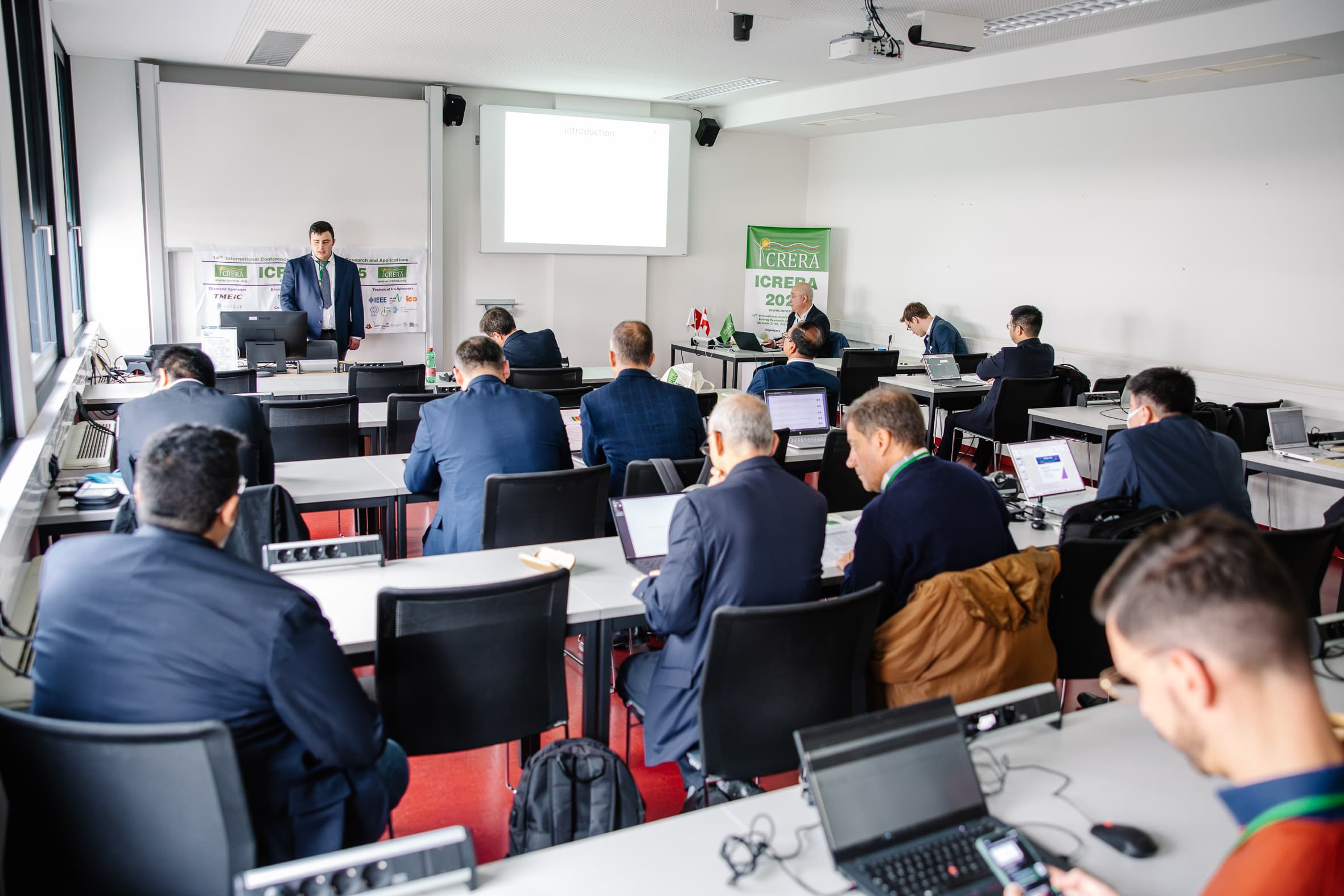 Eine Gruppe von Personen in Geschäftskleidung sitzt an Tischen mit Laptops in einem hellen Klassenzimmer und hört einem Redner zu, der an der Stirnseite neben einer Projektionsfläche und Konferenzbannern steht.