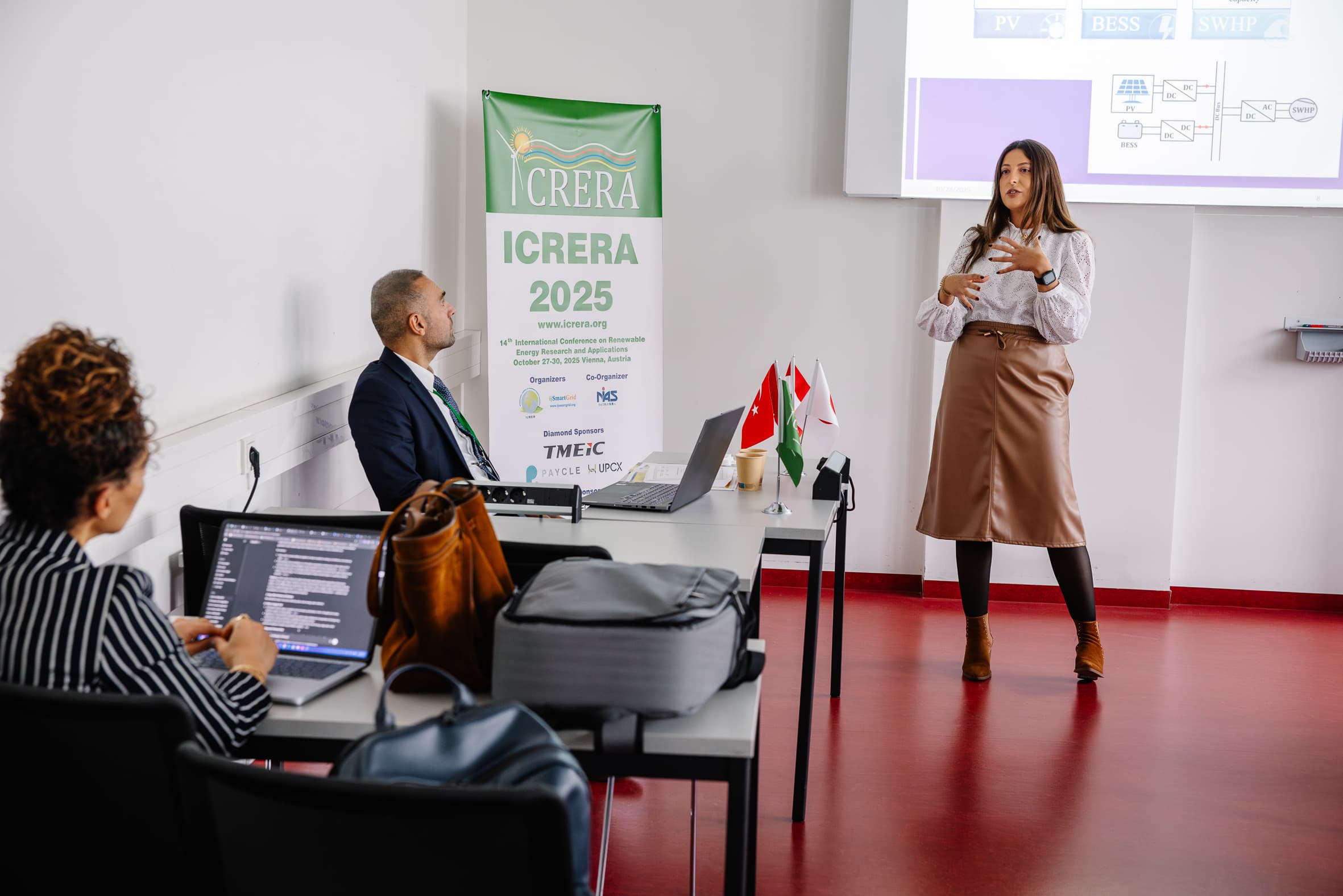 Eine Frau steht und präsentiert zwei sitzenden Kollegen in einem Konferenzraum, während auf dem Tisch Laptops geöffnet sind und im Hintergrund ein Veranstaltungsbanner der ICRERA 2025 hängt.