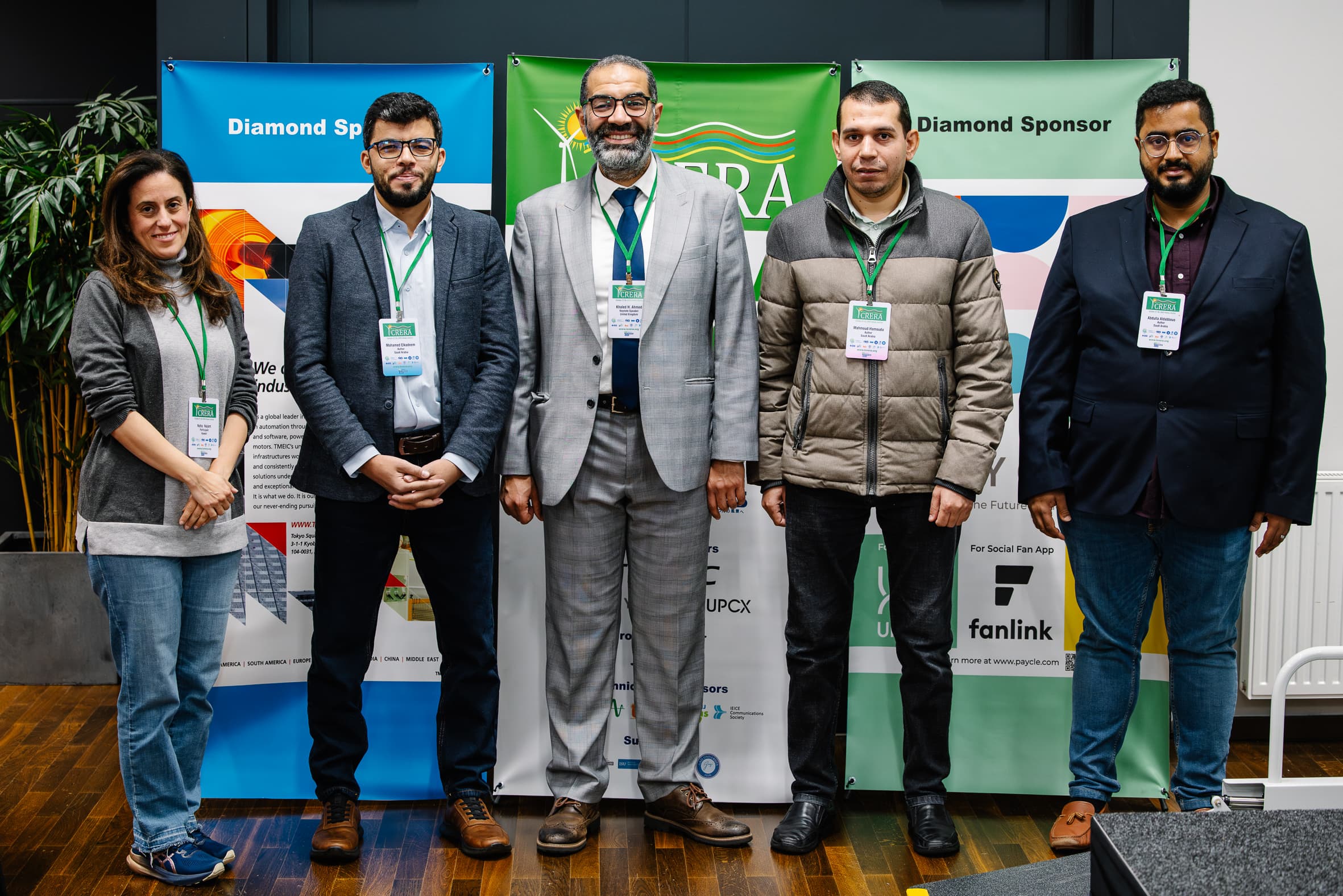 Fünf Personen stehen in einem Innenraum vor Sponsorenbannern bei einer Veranstaltung. Sie sind der Kamera zugewandt, tragen Namensschilder und legere Geschäftskleidung und scheinen an einer professionellen Versammlung oder Konferenz teilzunehmen.