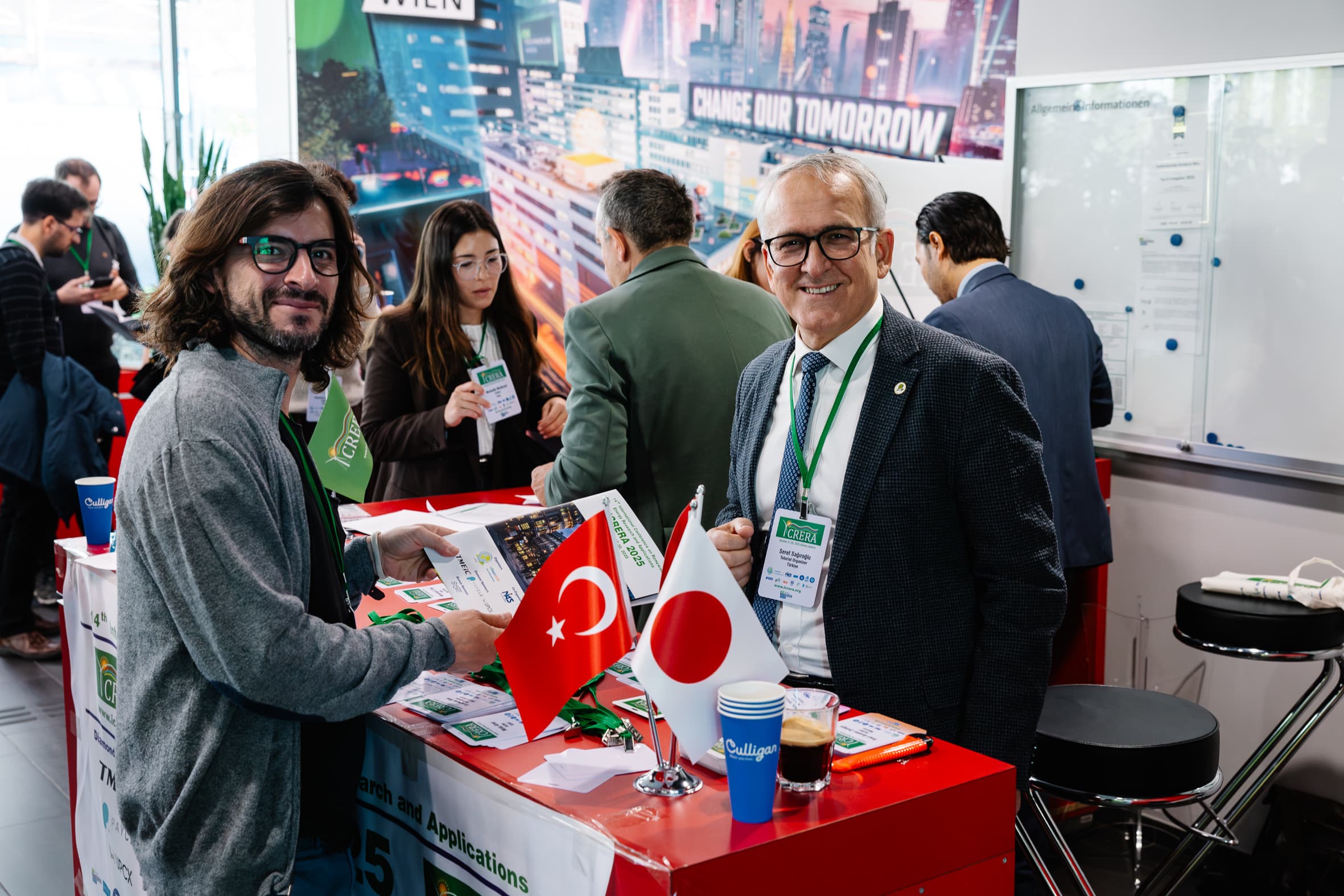 Zwei Männer stehen an einem Konferenzstand mit türkischen und japanischen Flaggen und lächeln in die Kamera. Im Hintergrund netzwerken andere Personen. Auf dem roten Tisch sind Broschüren, Anstecker und eine Kaffeetasse zu sehen.