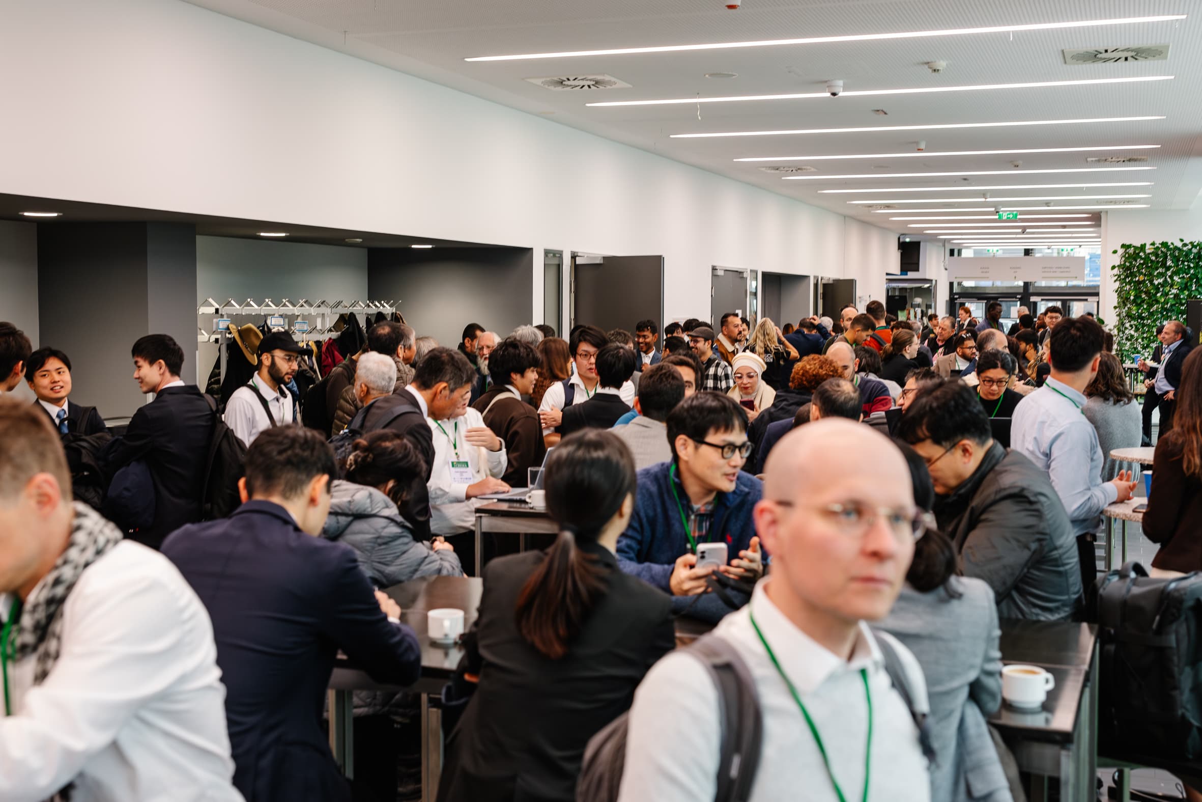 Eine große Gruppe von Menschen, viele in Geschäftskleidung, versammelt sich in einem hellen, modernen Flur, unterhält sich und knüpft Kontakte an Stehtischen während einer Konferenz oder einer beruflichen Veranstaltung.