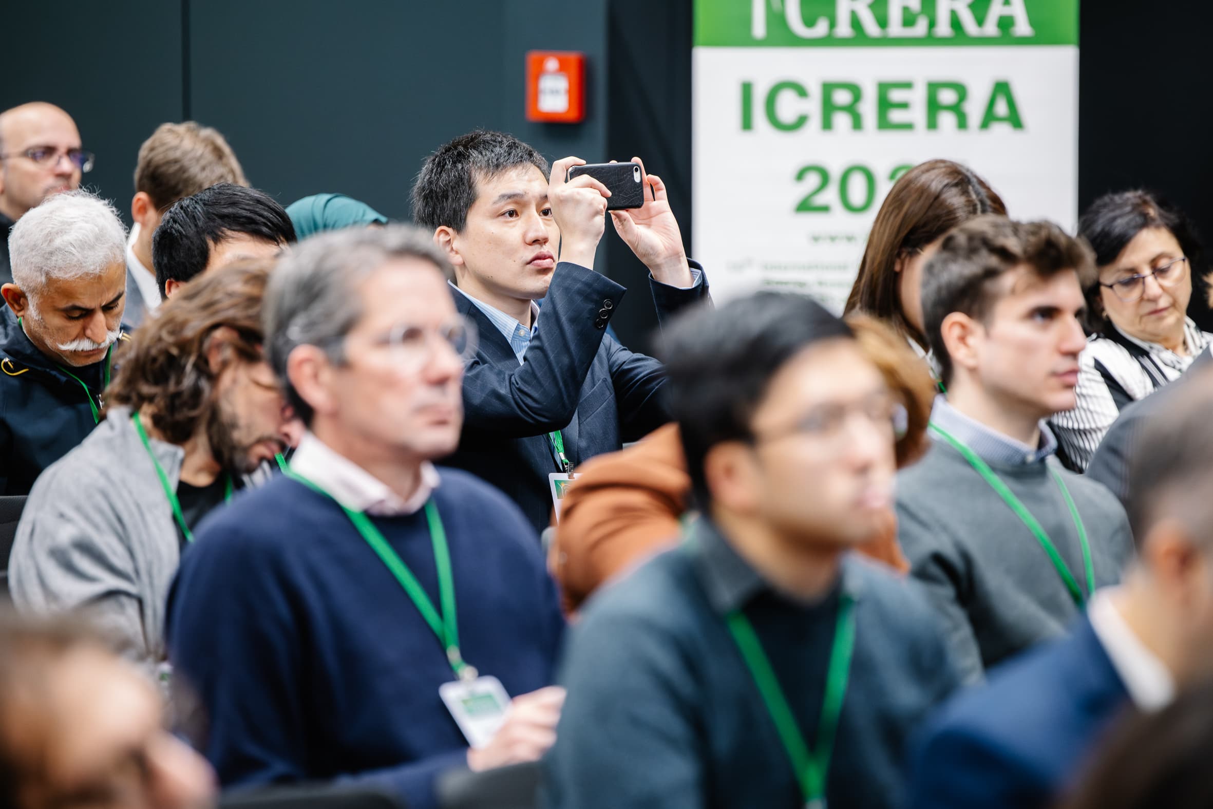 Eine bunt gemischte Gruppe von Menschen sitzt aufmerksam bei einer Konferenz; ein Mann im Anzug steht auf, um ein Foto mit seinem Handy zu machen. Im Hintergrund ist ein großes IC RERA 2021-Plakat zu sehen.