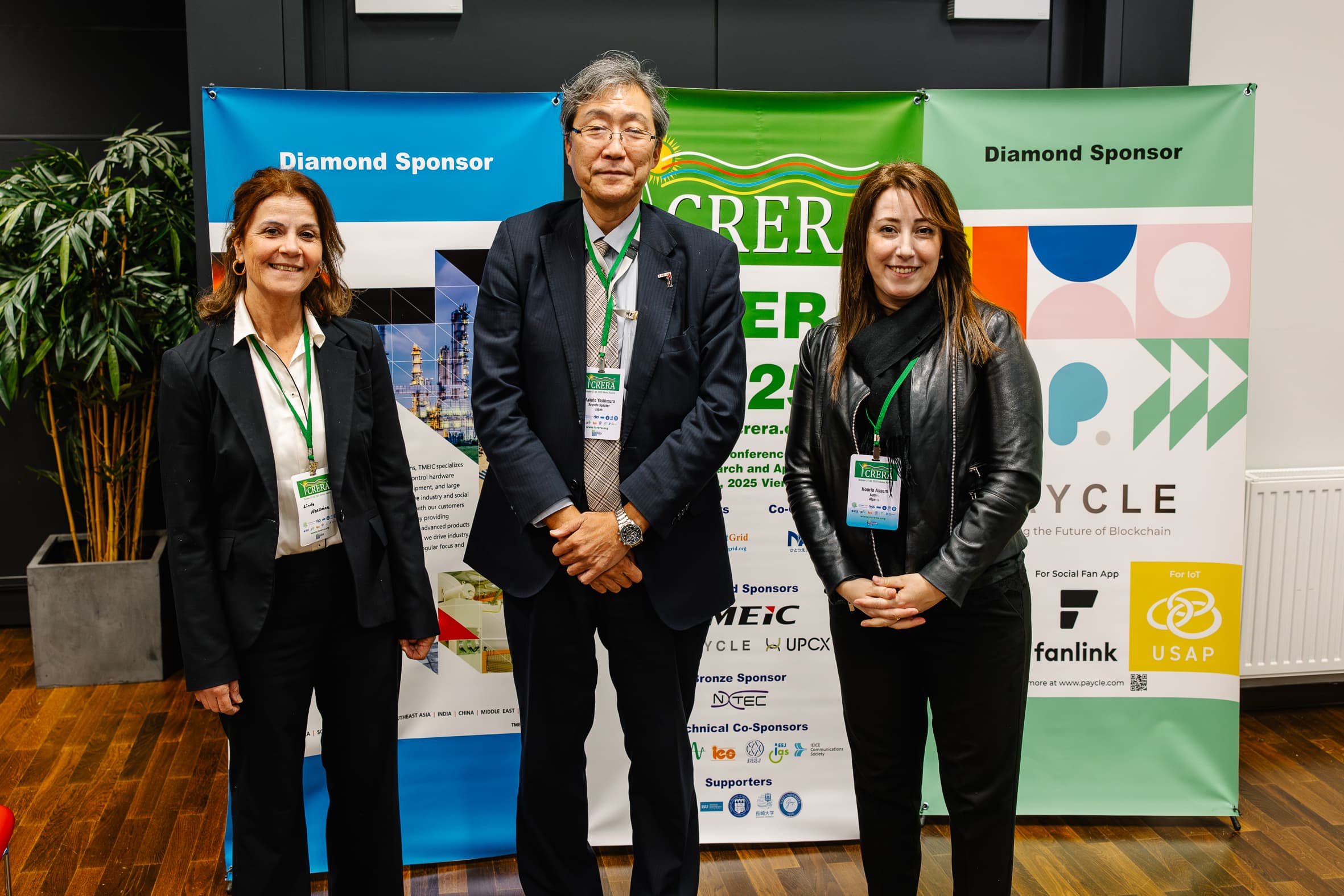Drei Personen mit Konferenzabzeichen stehen vor bunten Sponsorenbannern bei einer Veranstaltung. Zwei Frauen und ein Mann lächeln, gekleidet in Geschäftskleidung, mit Holzfußboden und einer Pflanze im Hintergrund.
