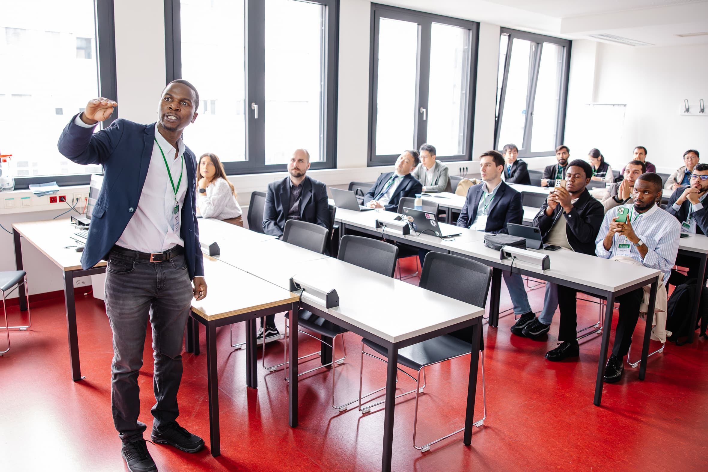 Ein Mann hält einen Vortrag vor einer Gruppe von aufmerksamen Zuhörern, die in einem modernen, gut beleuchteten Klassenzimmer mit großen Fenstern an Tischen sitzen. Einige Teilnehmer machen sich Notizen oder verwenden Laptops.