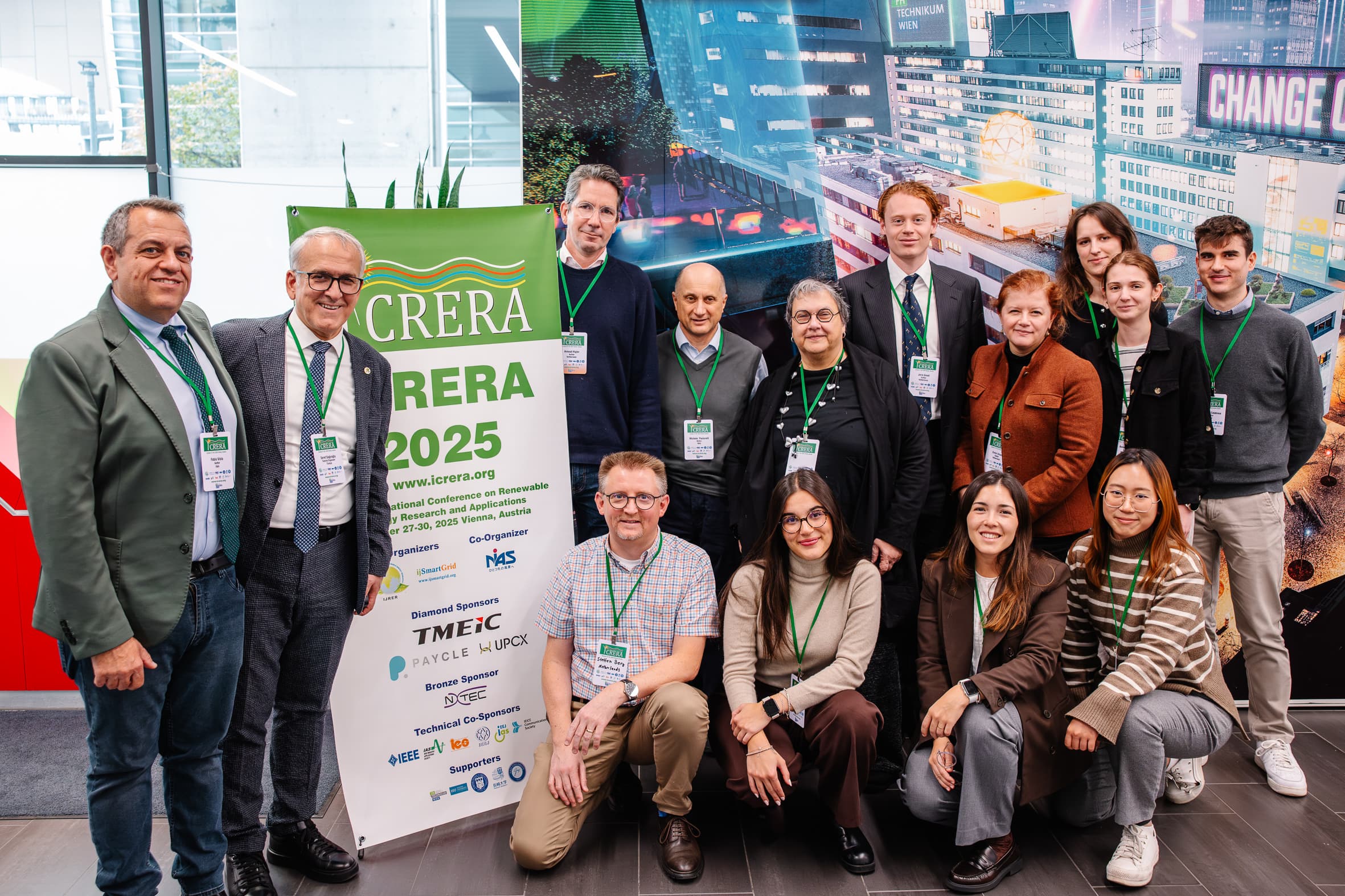 Eine Gruppe von fünfzehn Personen mit Konferenzabzeichen posiert drinnen neben einem grün-weißen Banner für LCRERA 2025. Einige stehen, andere knien davor, alle lächeln. Im Hintergrund ist ein städtisches Wandgemälde zu sehen.