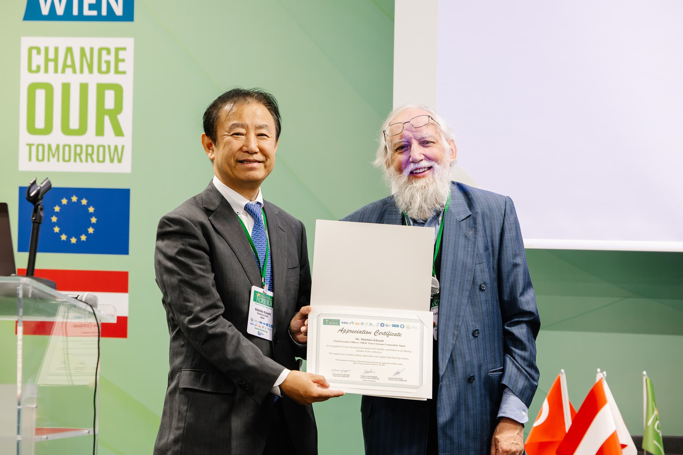Zwei Männer in Anzügen stehen lächelnd beieinander und halten bei einer Veranstaltung eine Dankesurkunde in der Hand. Hinter ihnen sind Transparente zu sehen, darunter "CHANGE OUR TOMORROW", und im Vordergrund sind mehrere Landesflaggen zu sehen.