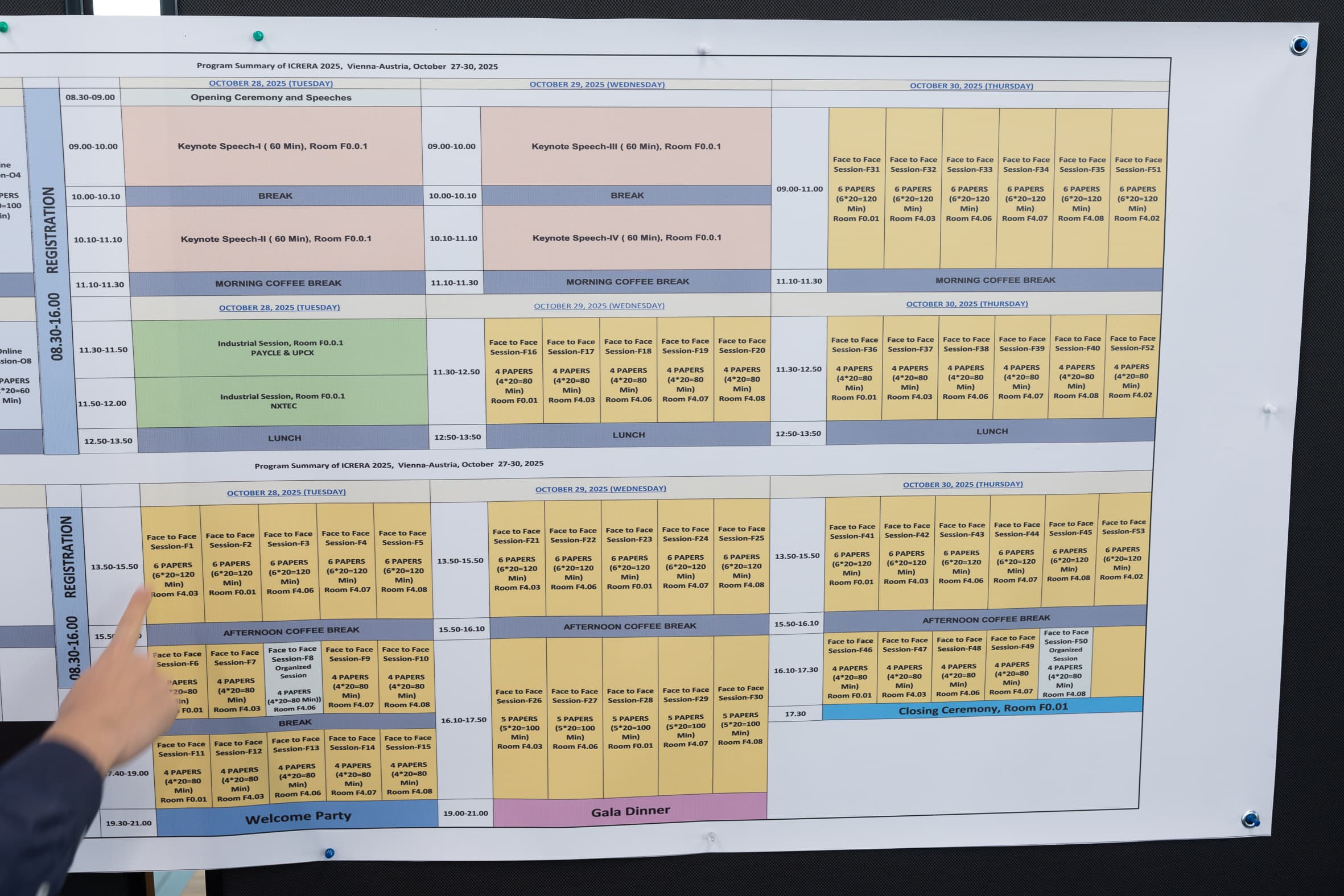 Eine Person zeigt auf einen detaillierten, farblich gekennzeichneten Veranstaltungsplan, der an einer Wand hängt. Der Zeitplan listet die Veranstaltungszeiten, Räume und Titel der Sitzungen in Spalten und Zeilen auf, wobei Pausen, Mittagessen und Zeremonien hervorgehoben sind.