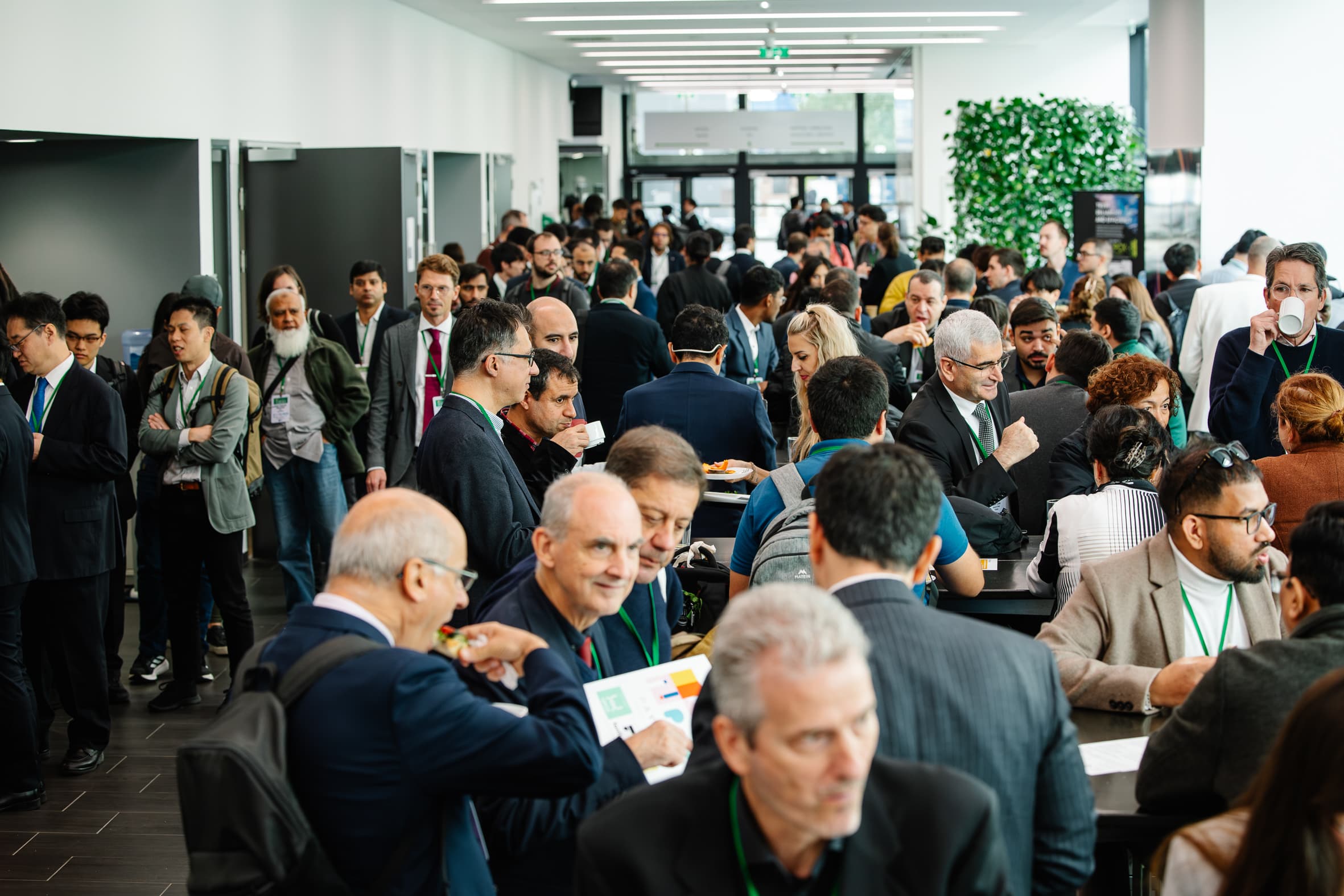 Eine große Gruppe von Menschen in Geschäftskleidung versammelt sich in einer hellen, modernen Lobby, mischt sich und knüpft Kontakte an Tischen während einer Konferenz oder einer beruflichen Veranstaltung.