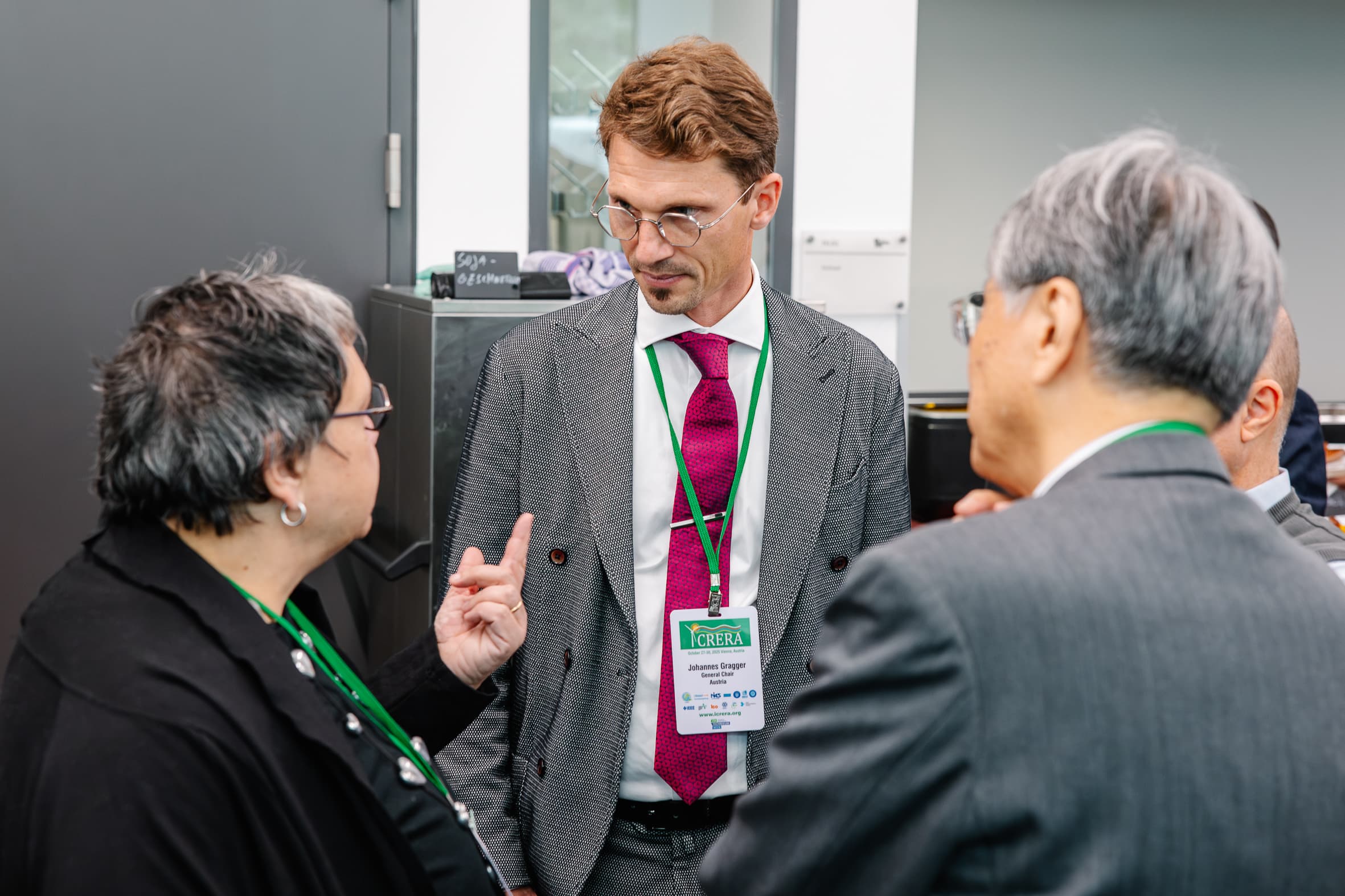 Drei Personen mit Konferenzschildern unterhalten sich in einem Innenraum. Eine Person gestikuliert mit der Hand, während sie mit einem Mann im Anzug und mit rosa Krawatte spricht; eine andere Person in einem grauen Anzug ist von hinten zu sehen.