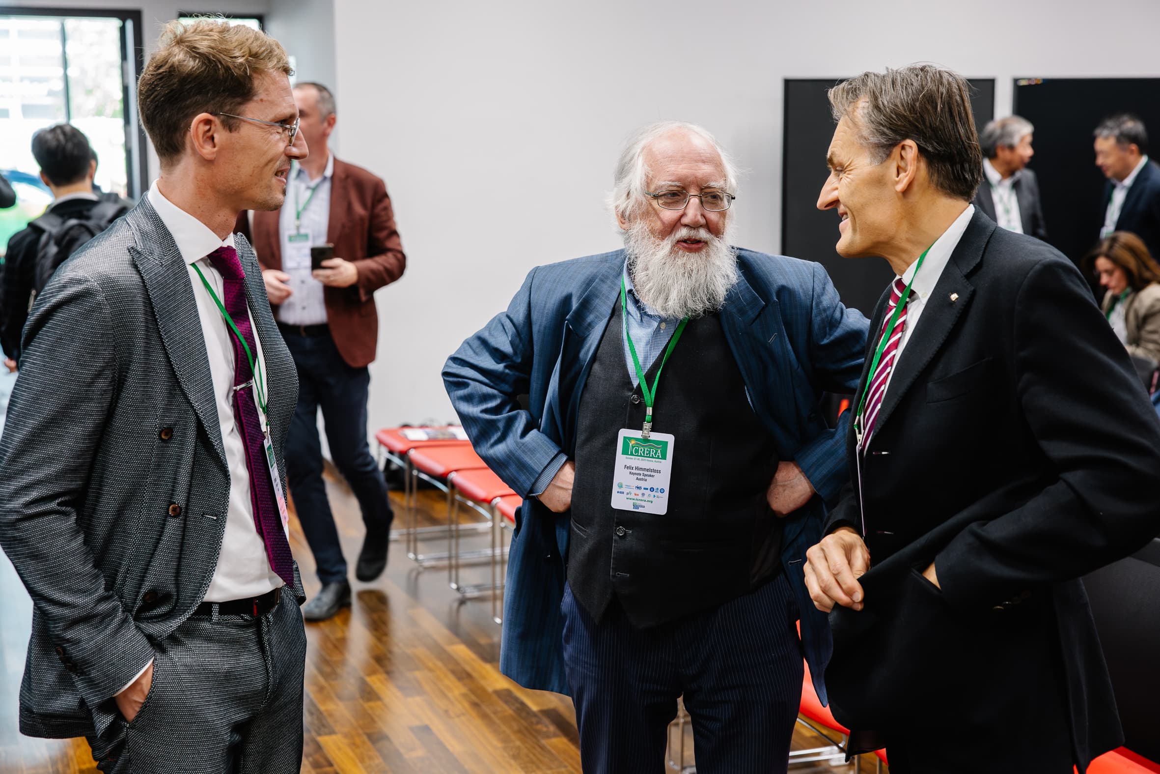 Drei Männer in formeller Kleidung stehen in einem Konferenzraum und unterhalten sich. Einer hat einen langen weißen Bart und trägt einen blauen Anzug. Sie tragen alle ein Konferenzabzeichen. Im Hintergrund sitzen andere Personen und unterhalten sich.