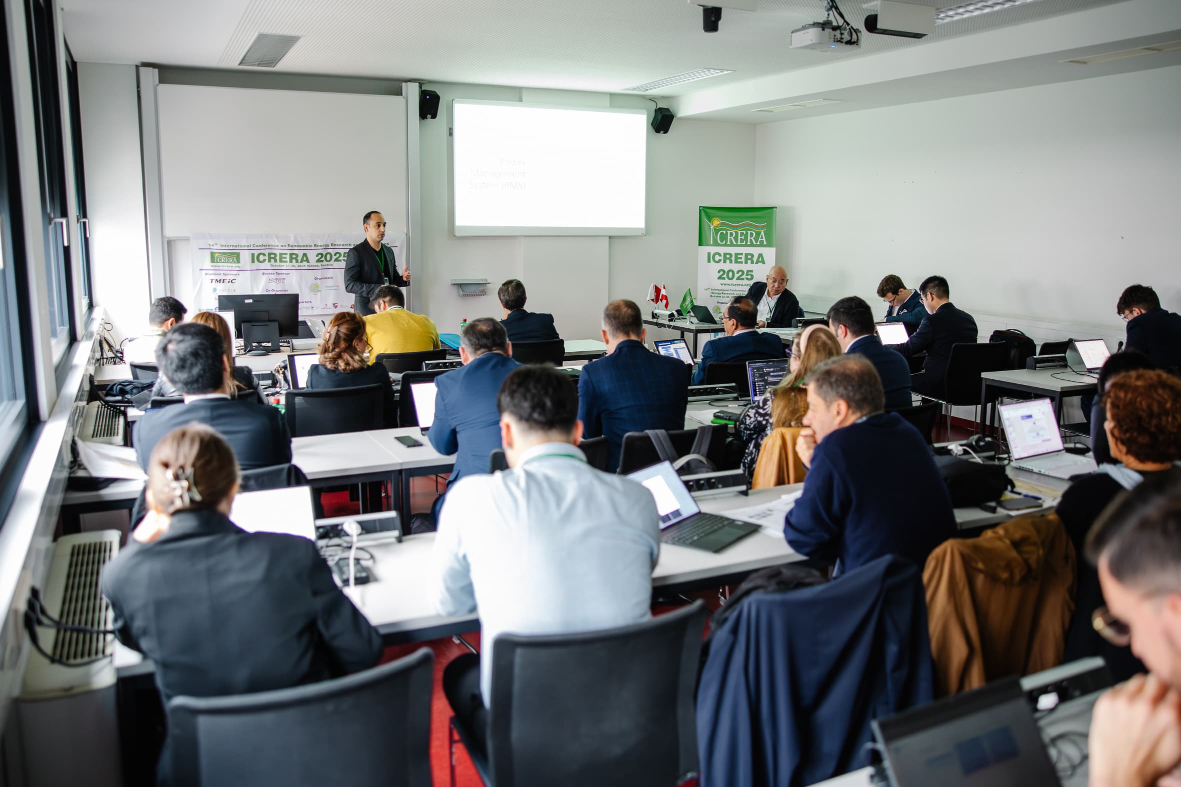 Eine Gruppe von Personen sitzt an Schreibtischen mit Laptops in einem hellen Konferenzraum und hört einem Vortragenden zu, der vorne neben einem Bildschirm steht, auf dem eine Präsentation gezeigt wird. Es sind Banner mit der Aufschrift "ICRERA 2025" zu sehen.