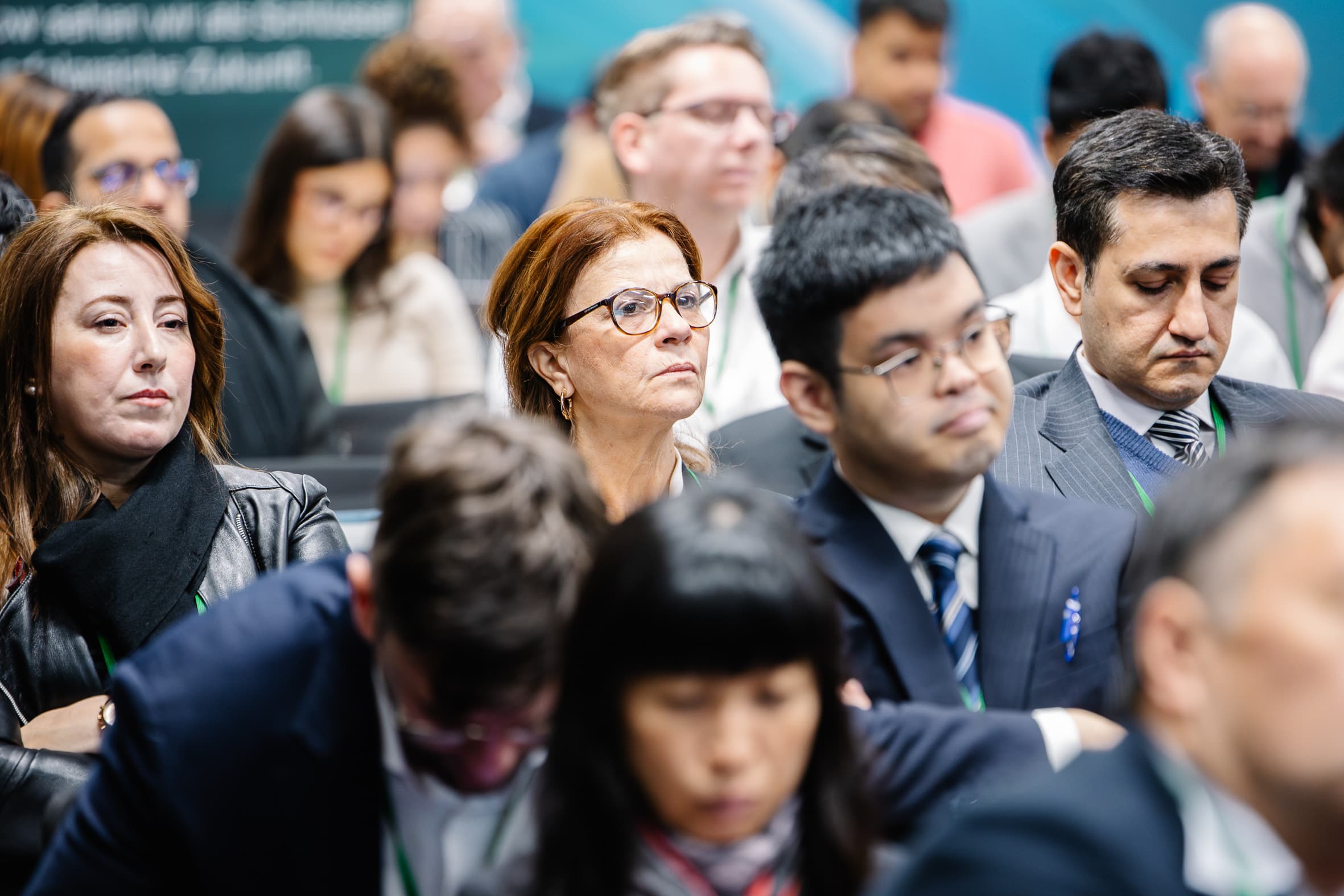 Eine bunt gemischte Gruppe von Menschen in Geschäftskleidung sitzt aufmerksam in einer Konferenz- oder Seminarumgebung und konzentriert sich auf einen Redner oder eine Präsentation, die nicht zu sehen ist. Einige wirken engagiert, während andere abgelenkt oder in Gedanken versunken aussehen.