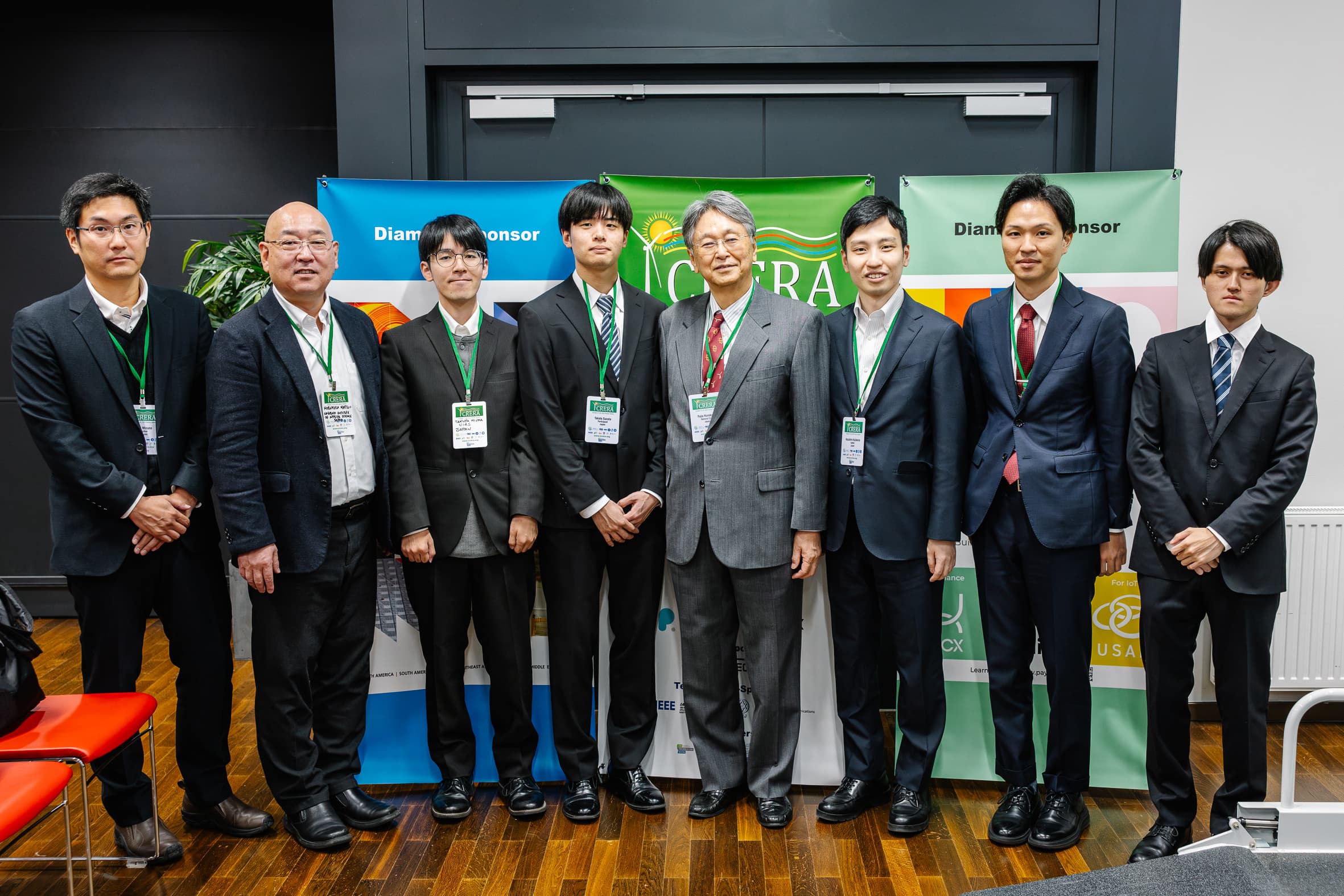 Acht Männer in Geschäftskleidung stehen Seite an Seite, der Kamera zugewandt, vor bunten Bannern und einer dunklen Wand. Die meisten tragen Anzüge und grüne Schlüsselbänder und posieren förmlich bei einer scheinbar professionellen Veranstaltung.
