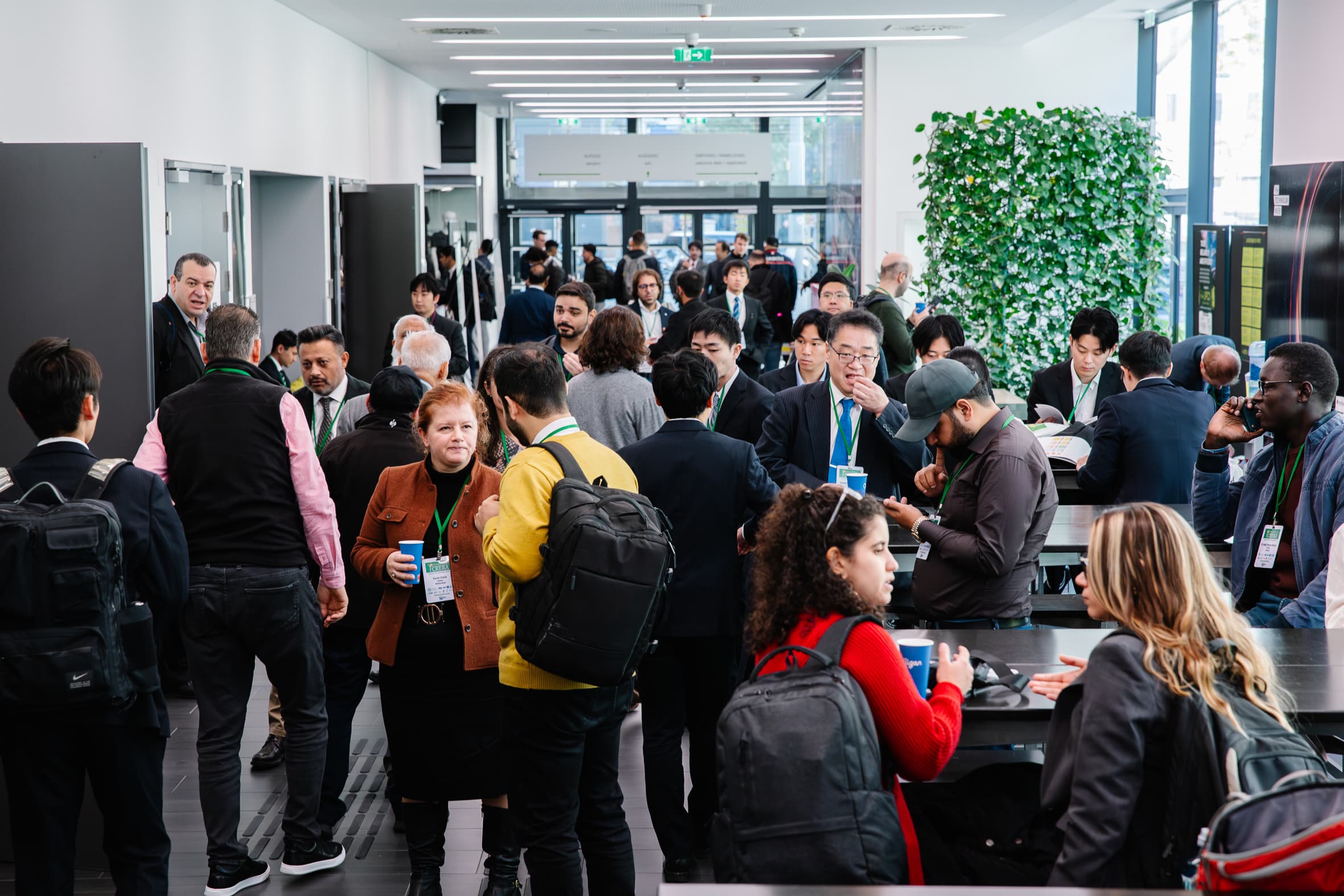 Eine große Gruppe von Menschen in Geschäftskleidung und mit Namensschildern versammelt sich in einem modernen, hell erleuchteten Raum mit Tischen, Pflanzen und Fenstern und unterhält sich. Einige sitzen, andere stehen und mischen sich.
