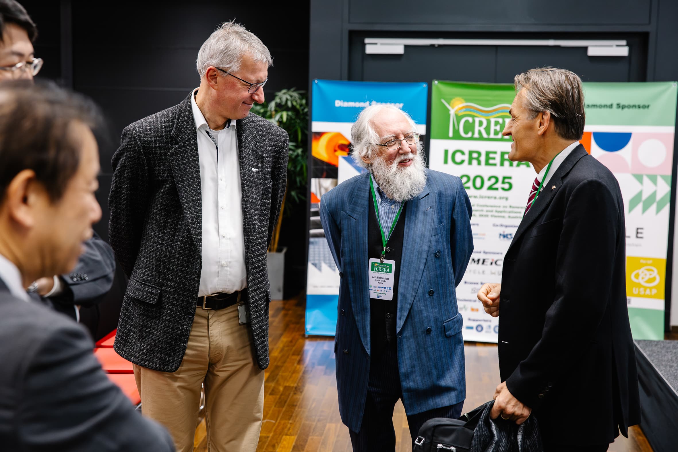 Eine Gruppe von fünf Männern in Geschäftskleidung unterhält sich angeregt auf einer Konferenz, während im Hintergrund Banner und Sponsorenlogos zu sehen sind. Ein Mann hält eine Jacke, ein anderer trägt ein Schlüsselband mit einem Konferenzausweis.
