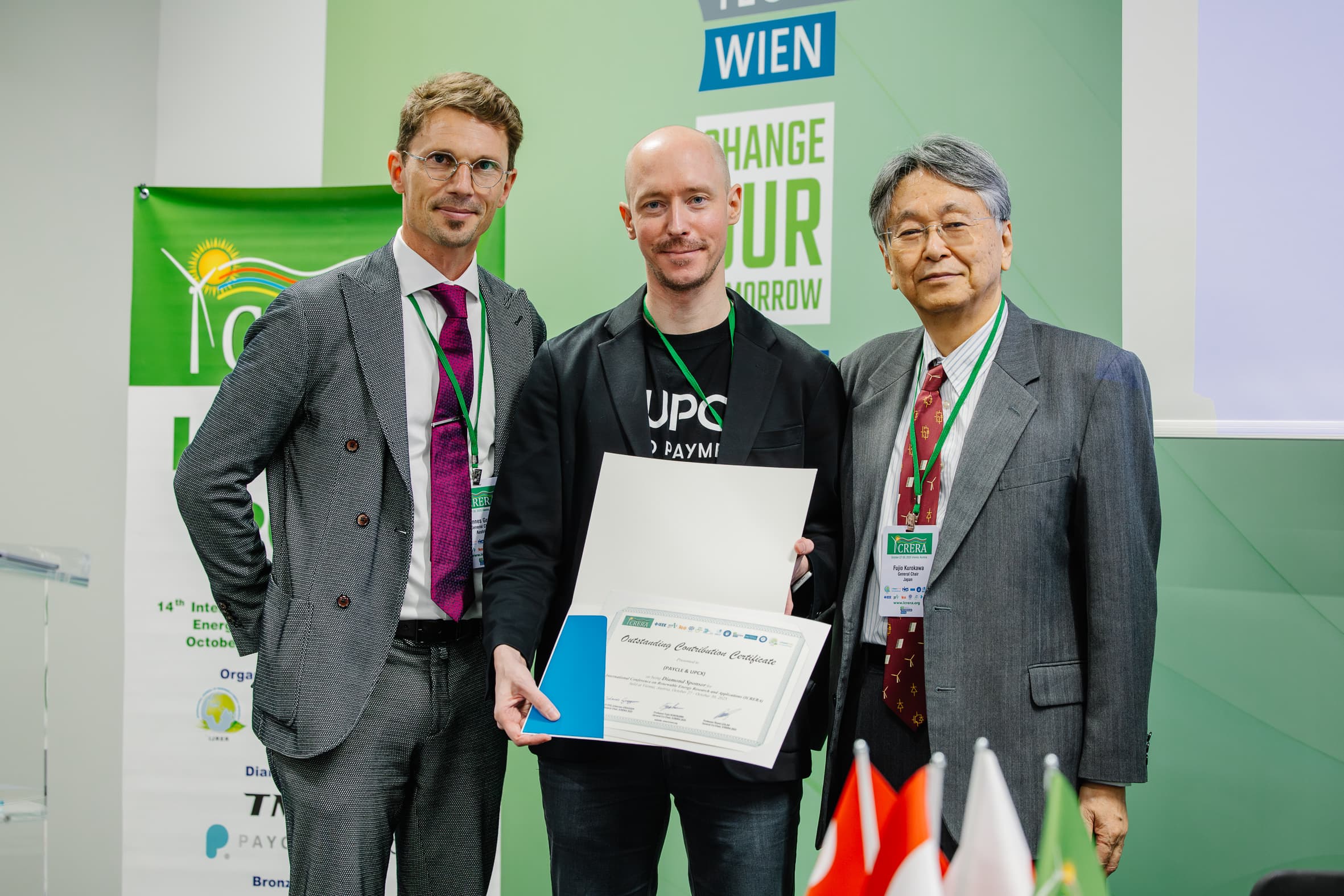 Drei Männer in Geschäftskleidung stehen bei einer Veranstaltung zusammen, einer hält eine Urkunde und lächelt. Hinter ihnen steht ein grünes Schild mit der Aufschrift WIEN und CHANGE YOUR TOMORROW. Im Vordergrund sind Fahnen und Veranstaltungsmaterialien zu sehen.