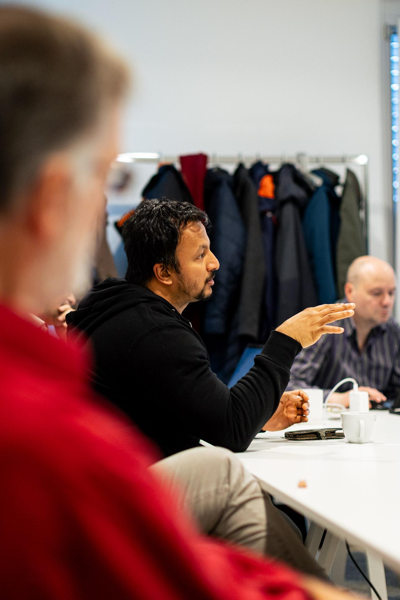 Ein Mann in einem schwarzen Kapuzenpulli gestikuliert, während er während einer Sitzung an einem Konferenztisch spricht. Im Hintergrund hängen Mäntel an einem Gestell, und in der Nähe sitzen weitere Personen, die teilweise unscharf sind.