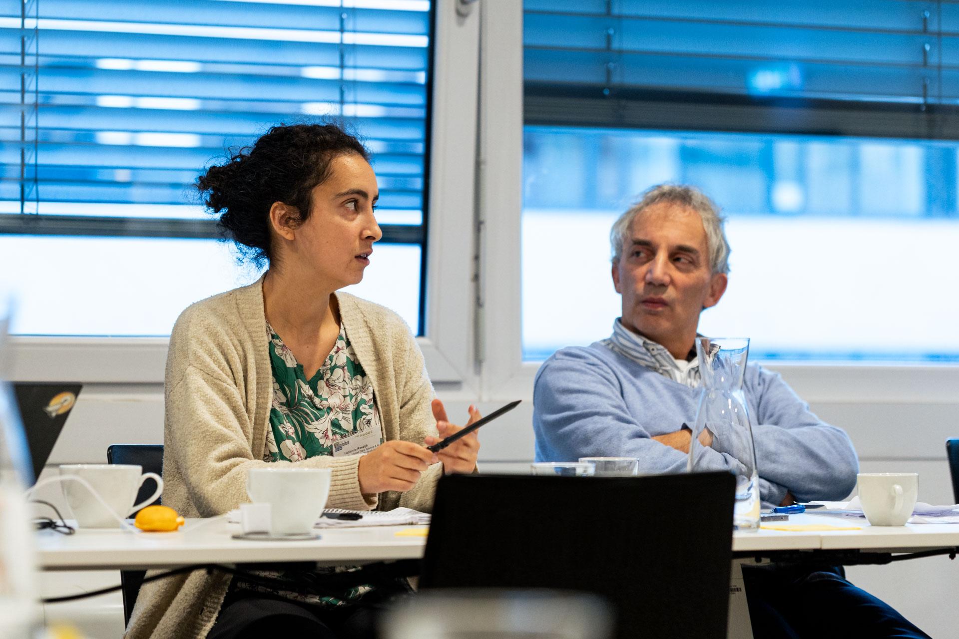 Eine Frau spricht während einer Besprechung und gestikuliert dabei mit einem Stift, während ein Mann neben ihr aufmerksam zuhört. Beide sitzen an einem Tisch mit Papieren, Tassen und einem Glaskrug in einem hellen Raum mit großen Fenstern.