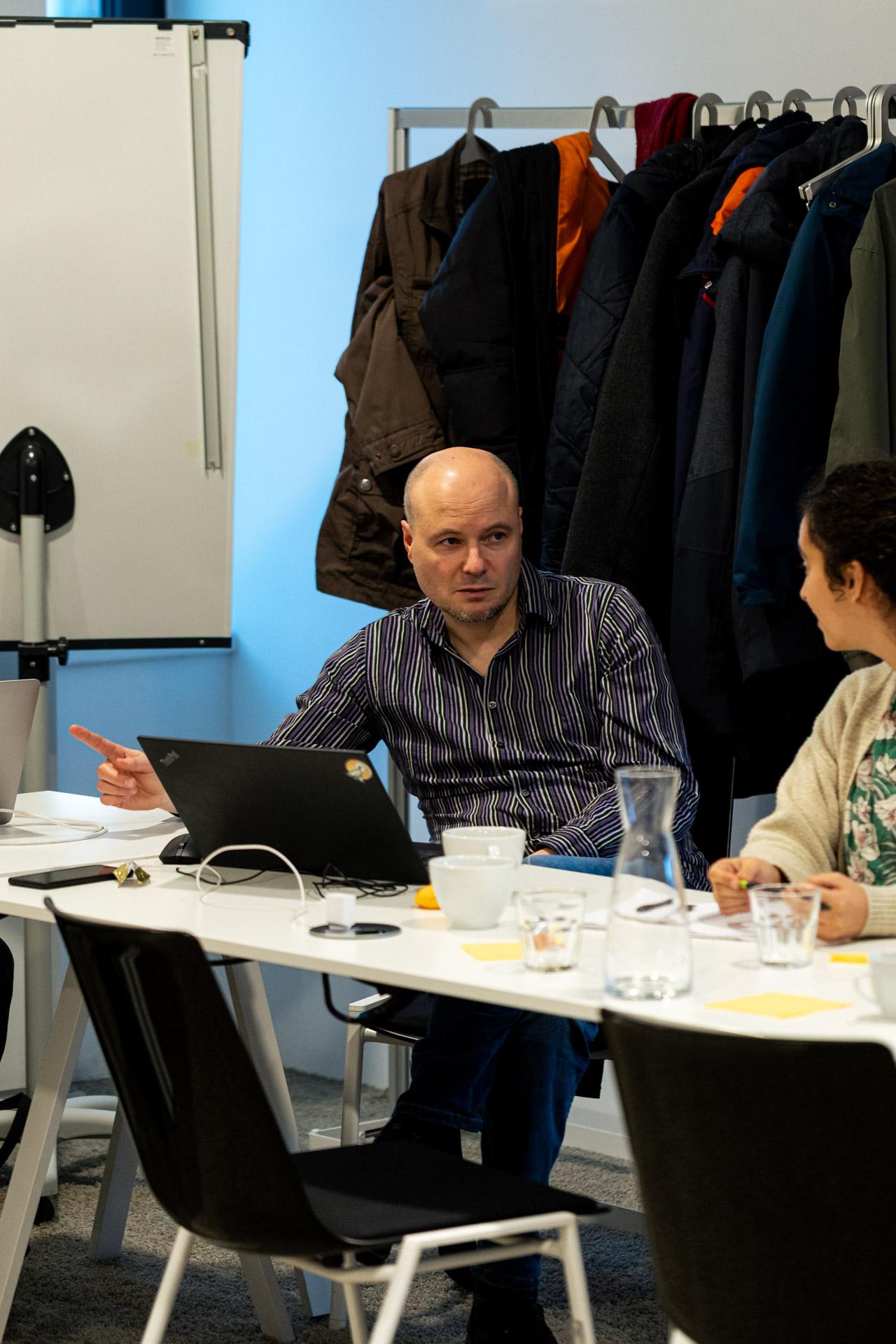 Ein Mann, der an einem Tisch mit einem Laptop sitzt, gestikuliert im Gespräch mit einer Frau. Hinter ihnen hängen Mäntel an einem Regal, und im Hintergrund ist ein Whiteboard zu sehen. Verschiedene Tassen und Wassergläser stehen auf dem Tisch.
