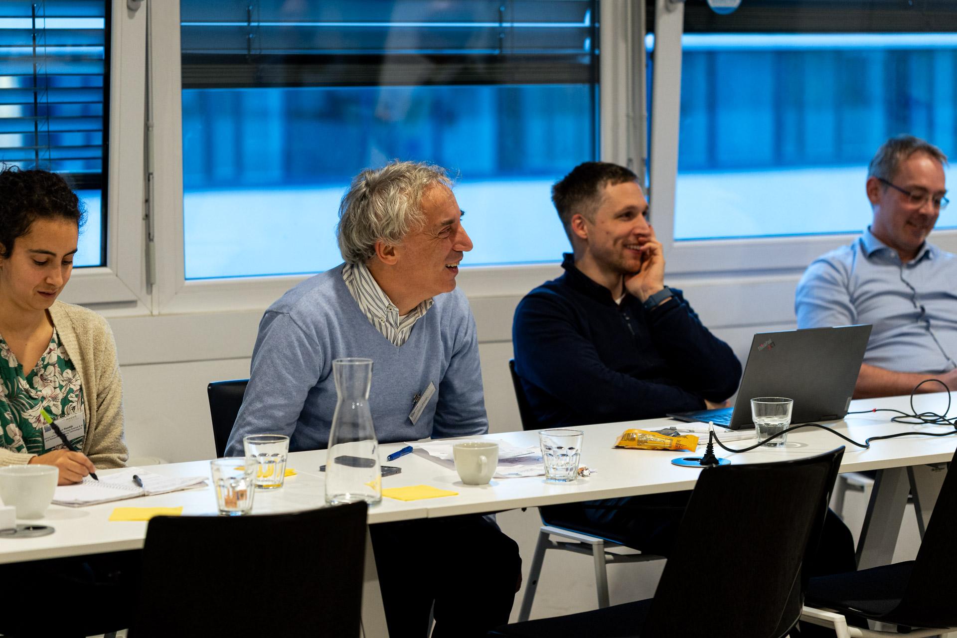 Vier Personen sitzen während einer Besprechung in einem hellen Raum an einem Tisch. Sie sind beschäftigt und lächeln, vor ihnen liegen Papiere, Getränke und ein Laptop. Große Fenster lassen natürliches Licht herein.