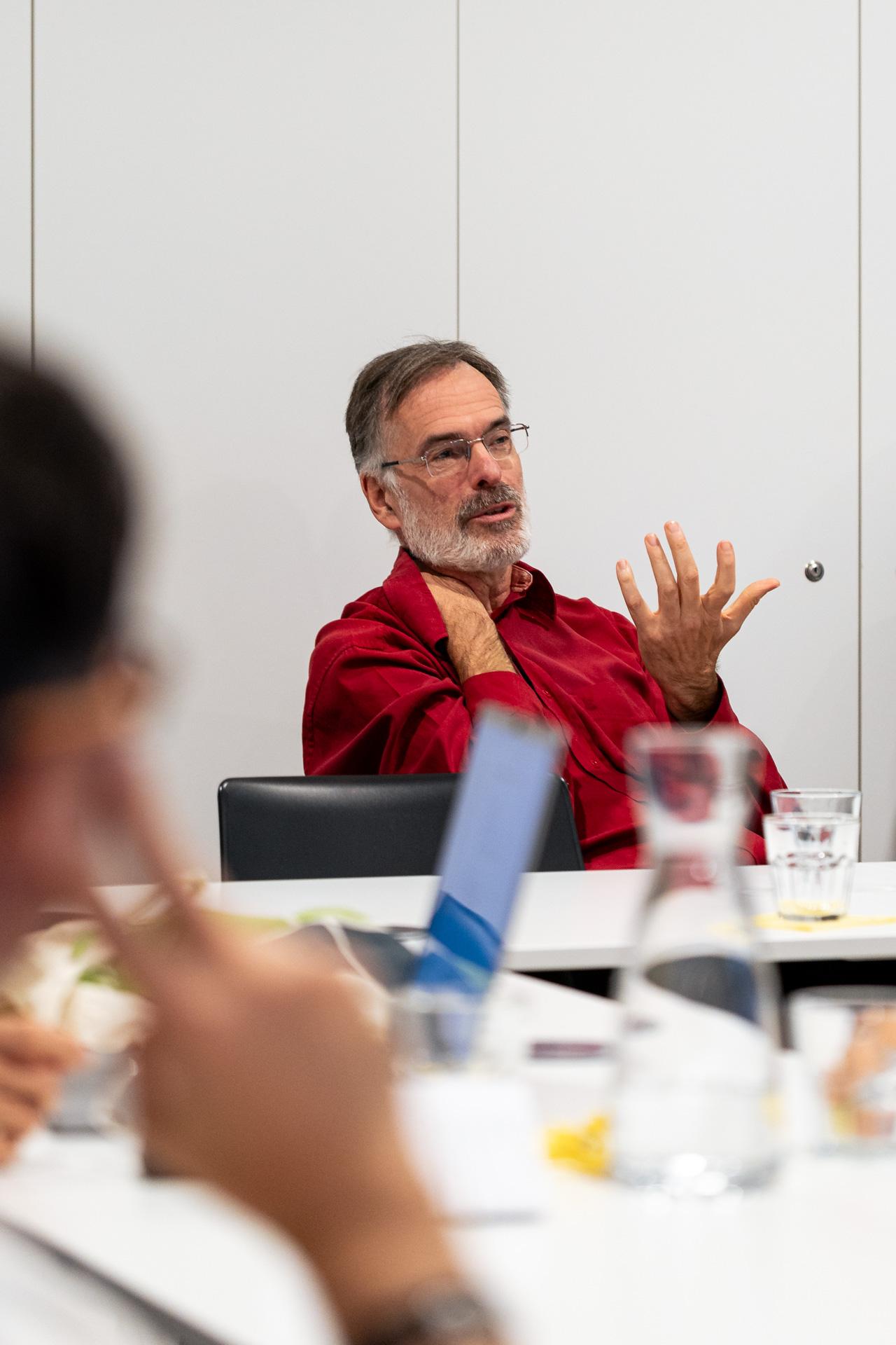 Ein Mann mit grauem Haar und Bart, der ein rotes Hemd trägt, sitzt an einem Tisch und gestikuliert mit einer Hand, während er mit der anderen seinen Nacken berührt. Er scheint in einer Besprechung oder Diskussion zu sprechen. Im Vordergrund sind unscharfe Menschen und ein Laptop zu sehen.