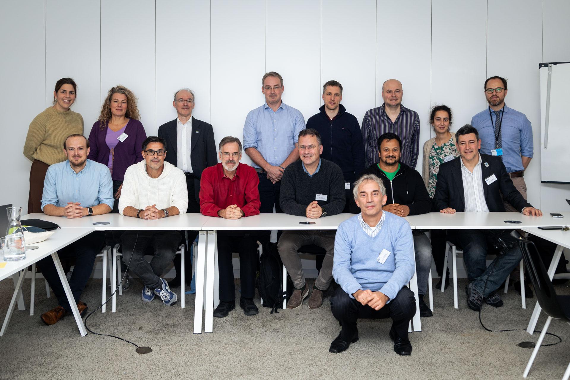 Fünfzehn Personen posieren für ein Gruppenfoto in einem Konferenzraum. Einige sitzen an einem weißen Tisch, andere stehen hinter ihnen. Sie sind in legerer Geschäftskleidung gekleidet und lächeln in die Kamera.