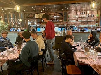 Eine Gruppe von Menschen sitzt um einen langen Tisch in einem gemütlichen Restaurant mit karierten Tischdecken, isst, spricht und telefoniert. Eine Person steht in der Mitte und unterhält sich mit den anderen am Tisch.