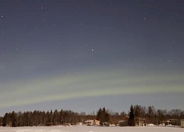Ein schwaches grünes Polarlicht erstreckt sich über einen sternenklaren Nachthimmel über einer verschneiten Landschaft mit Bäumen und einigen Häusern in der Nähe des Horizonts.