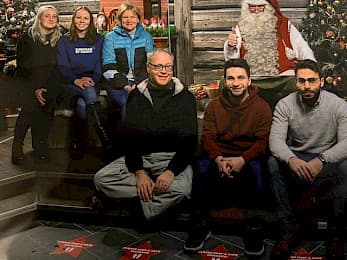 Eine Gruppe von sechs Personen, drei Männer und drei Frauen, sitzen und stehen vor einer festlichen Weihnachtsszene mit einem geschmückten Baum und einem als Weihnachtsmann verkleideten Mann, der im Hintergrund die Daumen nach oben zeigt.