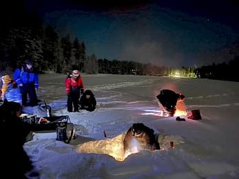 Eine Gruppe von Menschen in einer nächtlichen Schneelandschaft versammelt sich um Löcher im Schnee, die von Laternen beleuchtet werden. Im Hintergrund sind Bäume zu sehen, und in der Ferne leuchten Lichter, die eine heitere Winterszene schaffen.