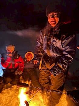 Drei in Winterkleidung gekleidete Personen sitzen nachts im Schnee, lächeln und wärmen sich an einem Lagerfeuer, an dem sie Essen auf Stöcken rösten. Die Szene ist durch das Feuer warm erleuchtet.