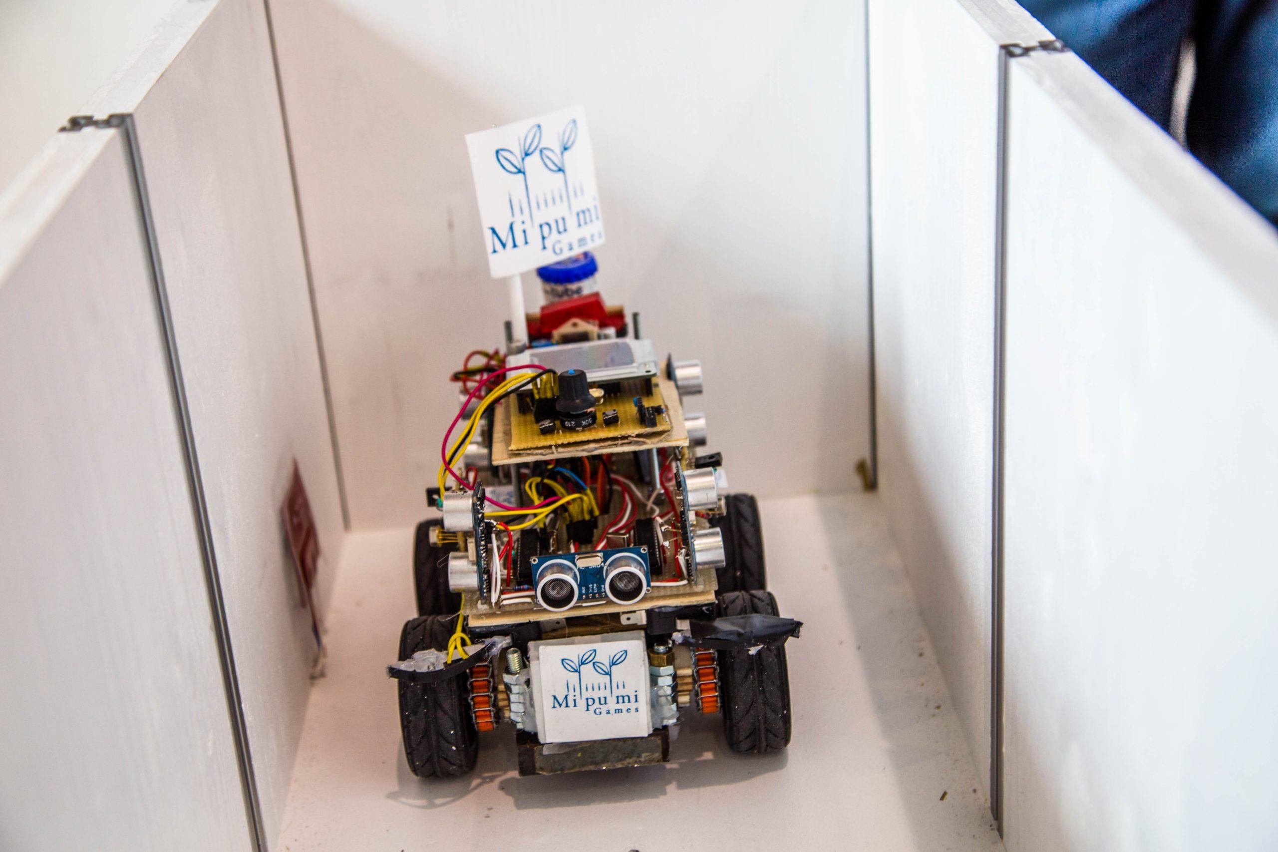 Ein kleiner Roboter auf Rädern mit freiliegenden elektronischen Komponenten und Drähten bewegt sich durch ein schmales weißes Gehäuse. An dem Roboter sind Banner mit der Aufschrift 'Mipumi Games' angebracht.