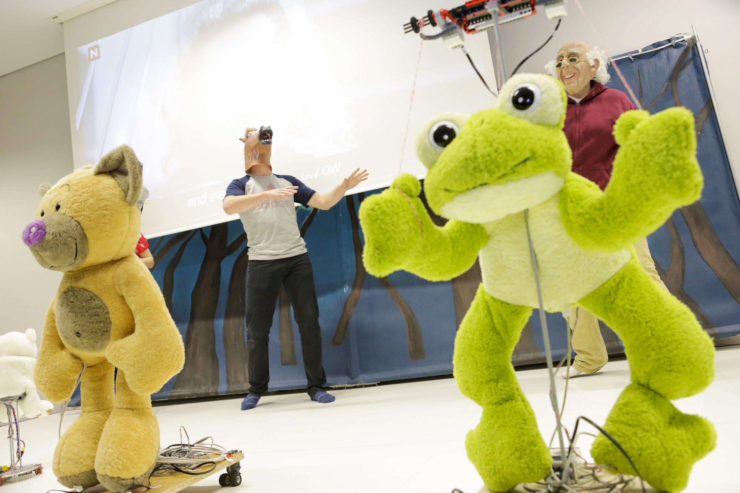 Zwei Personen mit Tiermasken treten hinter großen Plüschpuppen - einer Katze und einem Frosch - auf, die an Drähten und Mechanismen auf einer Bühne befestigt sind, mit einer projizierten Leinwand und einer Waldkulisse im Hintergrund.