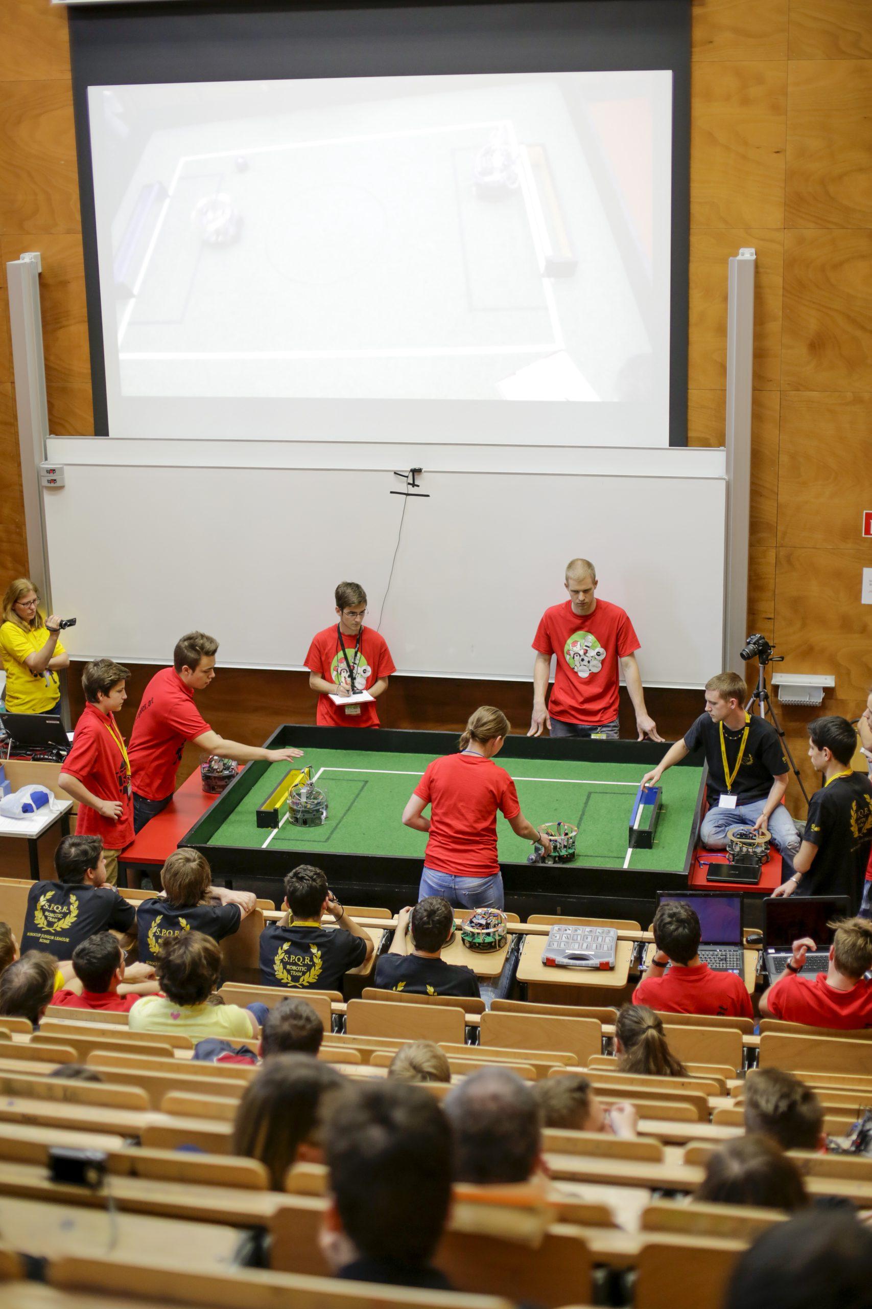 Eine Gruppe von Studenten in roten Hemden tritt in einem Robotikspiel an einem grünen Tisch vor einem Publikum in einem Hörsaal an, während hinter ihnen eine Projektion des Spiels auf einer Leinwand zu sehen ist.