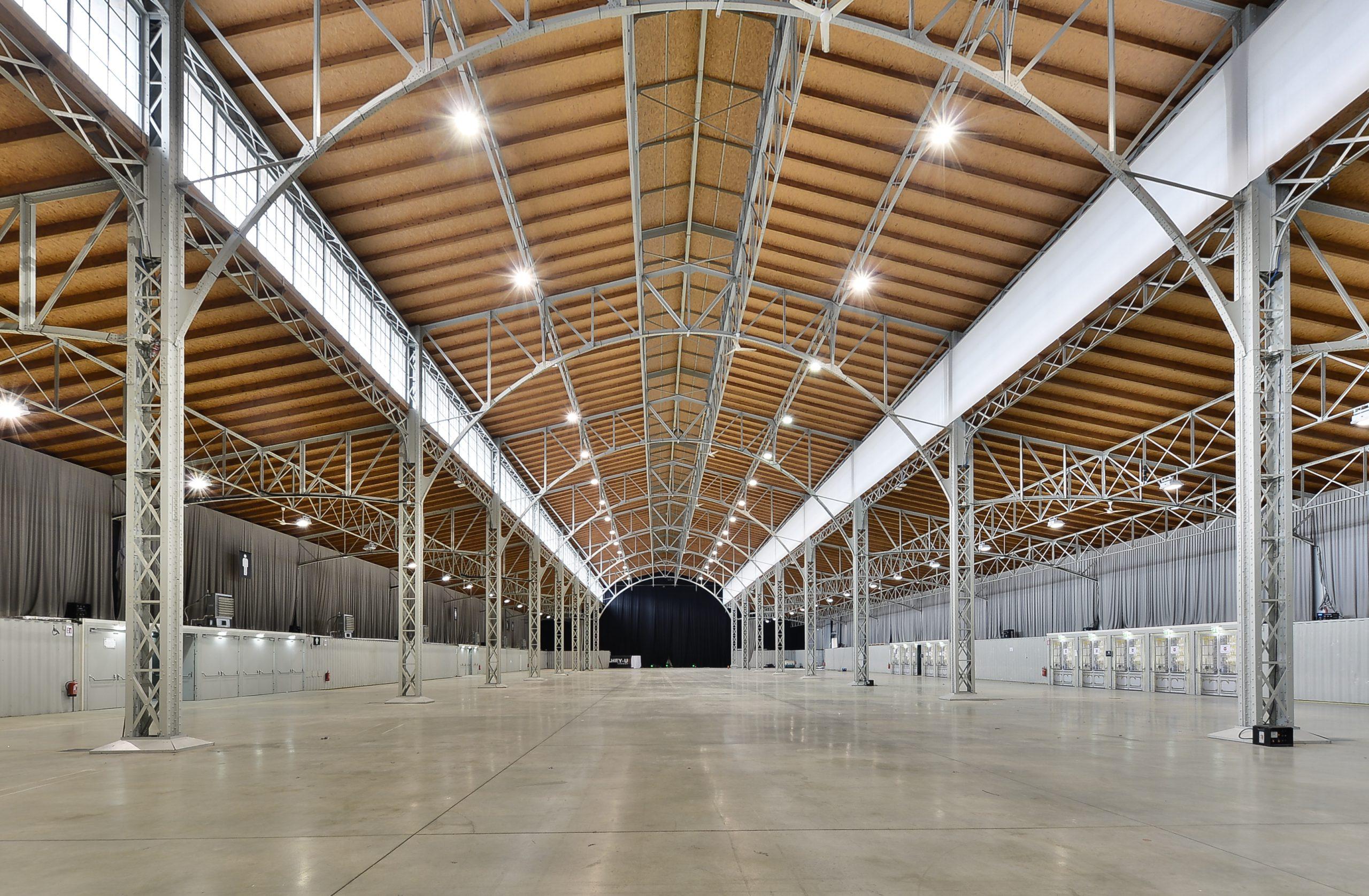 Geräumiges, leeres Industrielager mit hohen Gewölbedecken, freiliegenden Metallträgern, großen Fenstern und polierten Betonböden. Der Raum ist gut beleuchtet und symmetrisch, mit Vorhängen und weißen Trennwänden entlang der Wände.